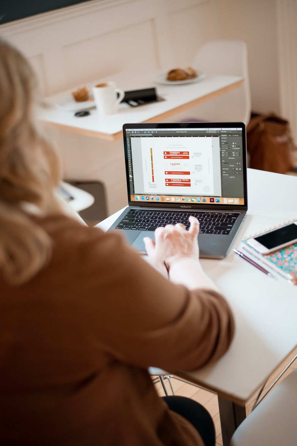 Person working on design software on laptop with coffee and pastry on white desk.