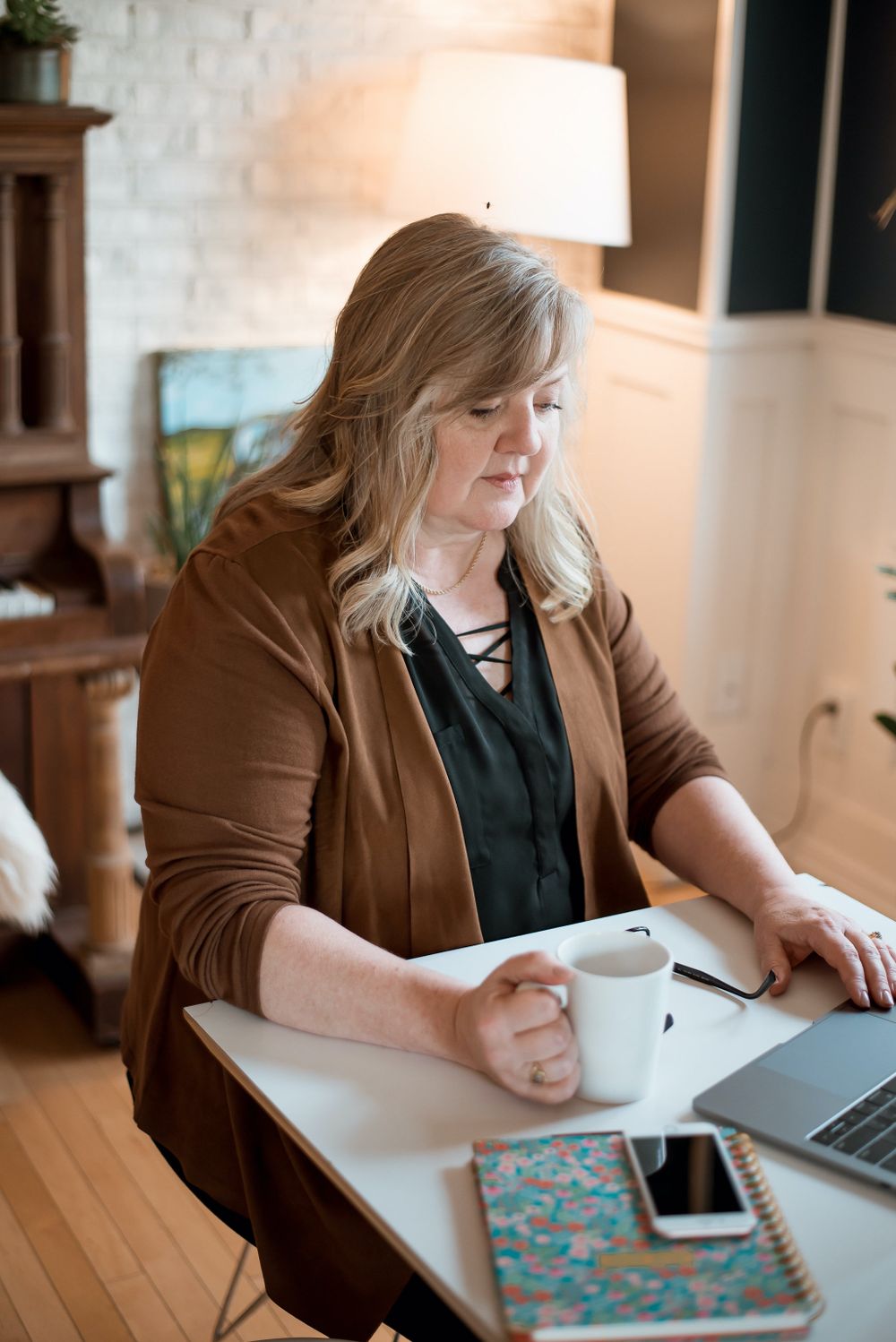 Side view of someone working at desk with laptop, coffee cup and floral notebook in warm light.