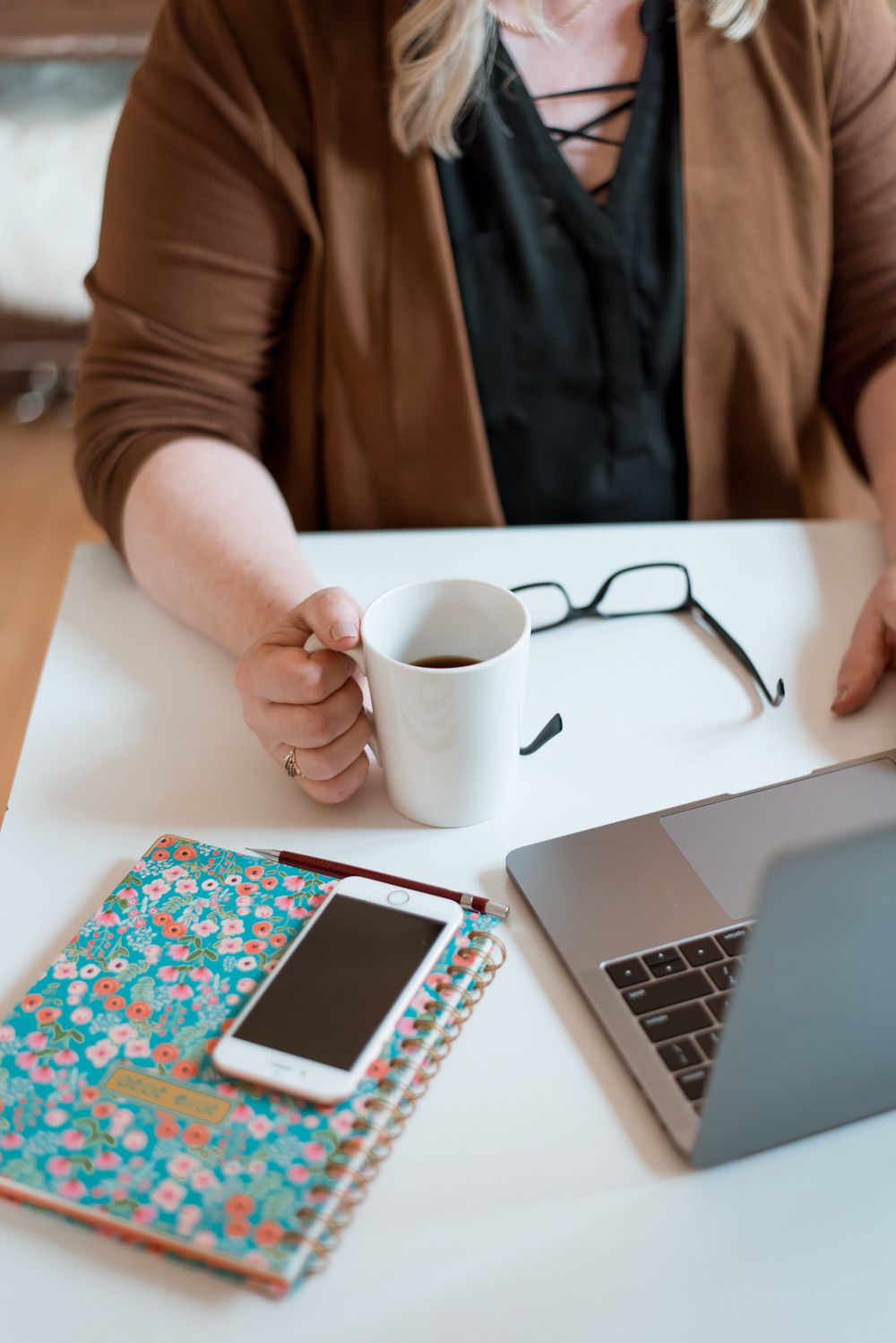 Desktop workspace with coffee mug, laptop, glasses and teal floral notebook on white surface.