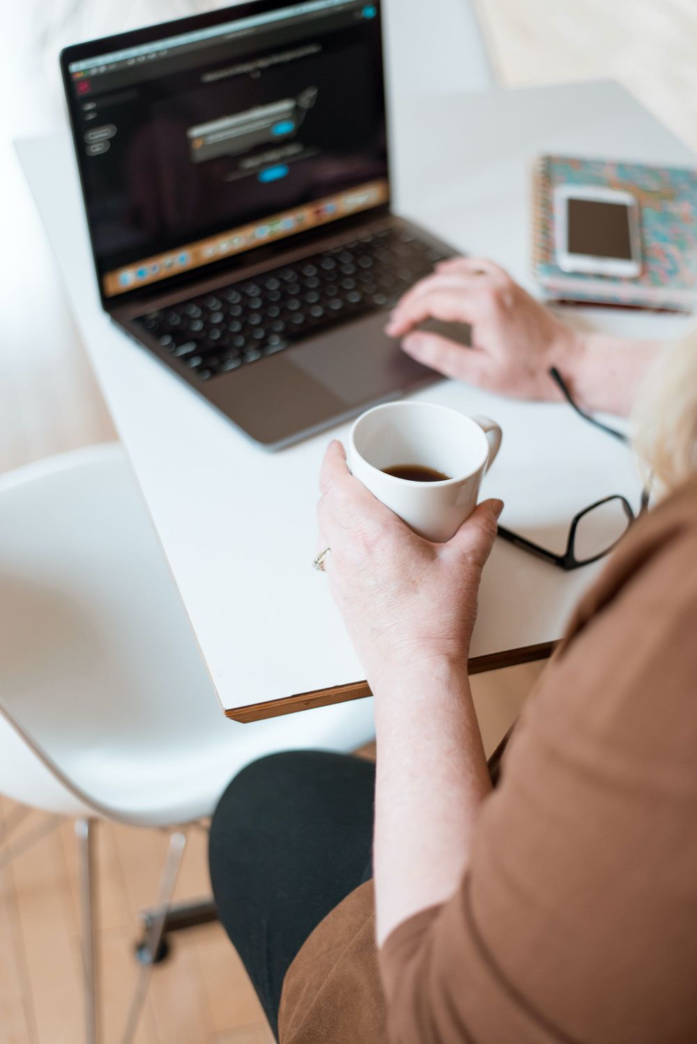 Hand holding coffee cup at workspace with laptop and glasses on white desk.