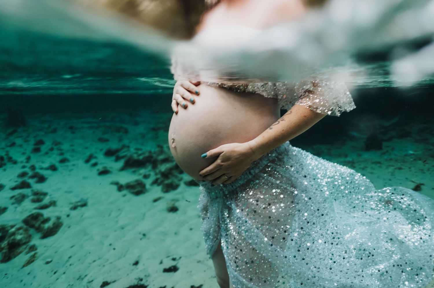 Underwater maternity photo showing a flowing dress and pregnant belly in turquoise water.