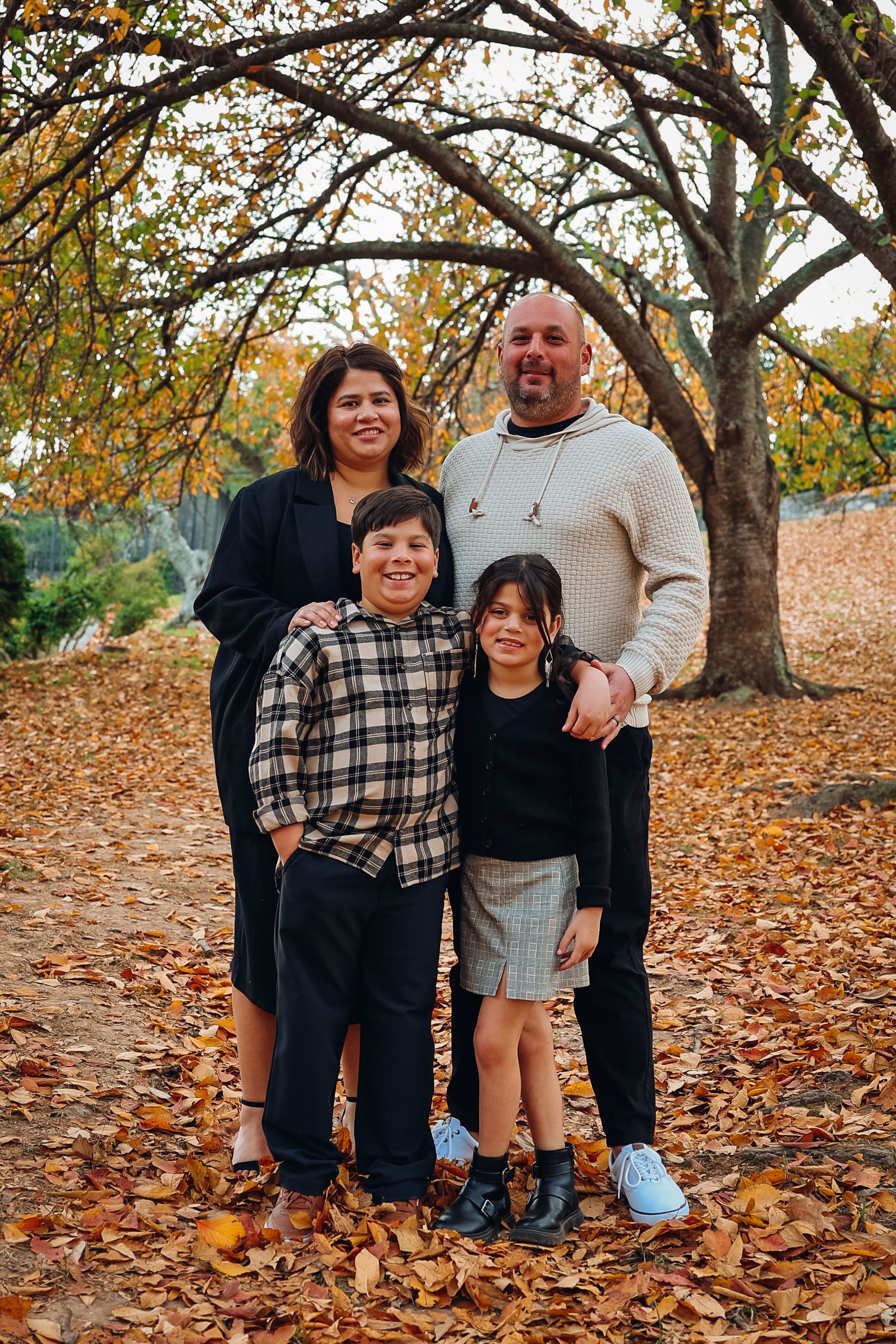 Family of four poses together on fallen autumn leaves under tree branches.