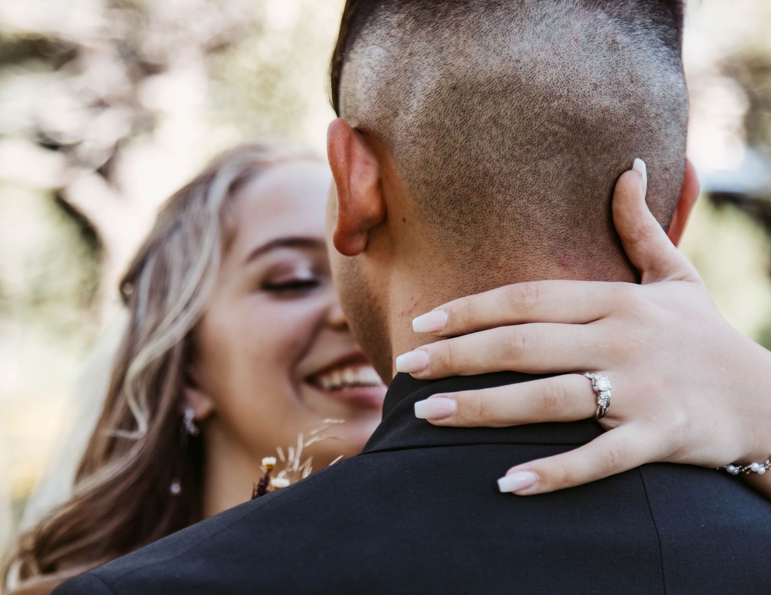 Close-up of an engagement ring shown as couple embraces outdoors in natural lighting.
