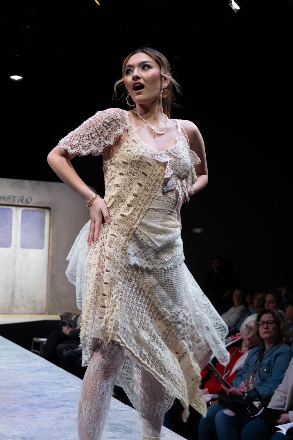 Model in cream lace asymmetrical dress walking the runway at a fashion show.