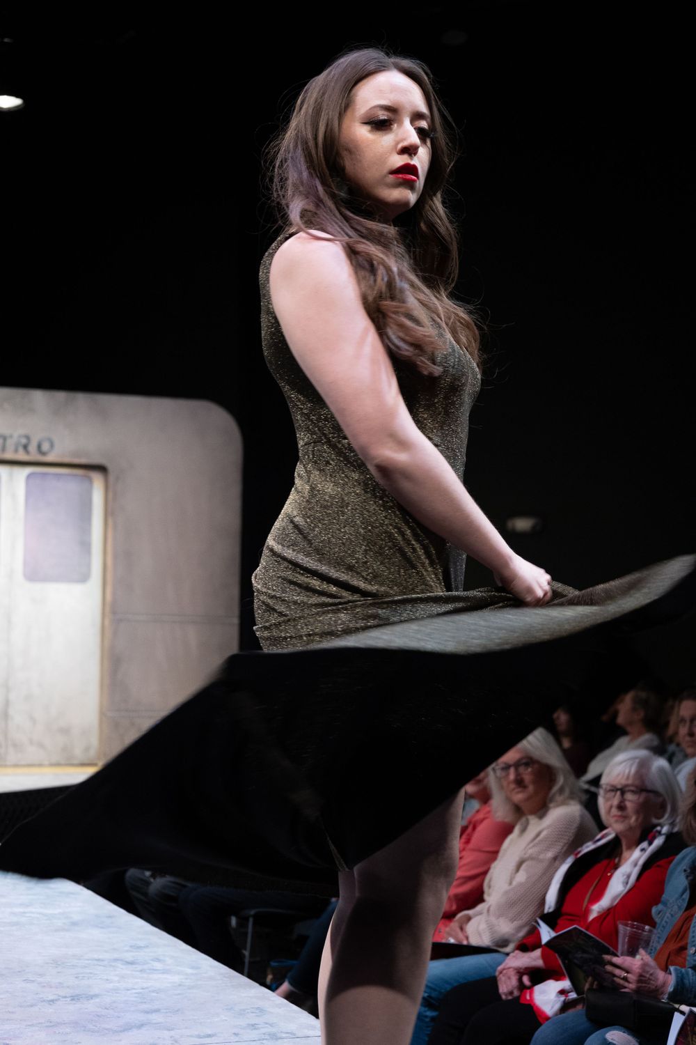 Model in olive green mini dress with black cape walking runway at fashion show.