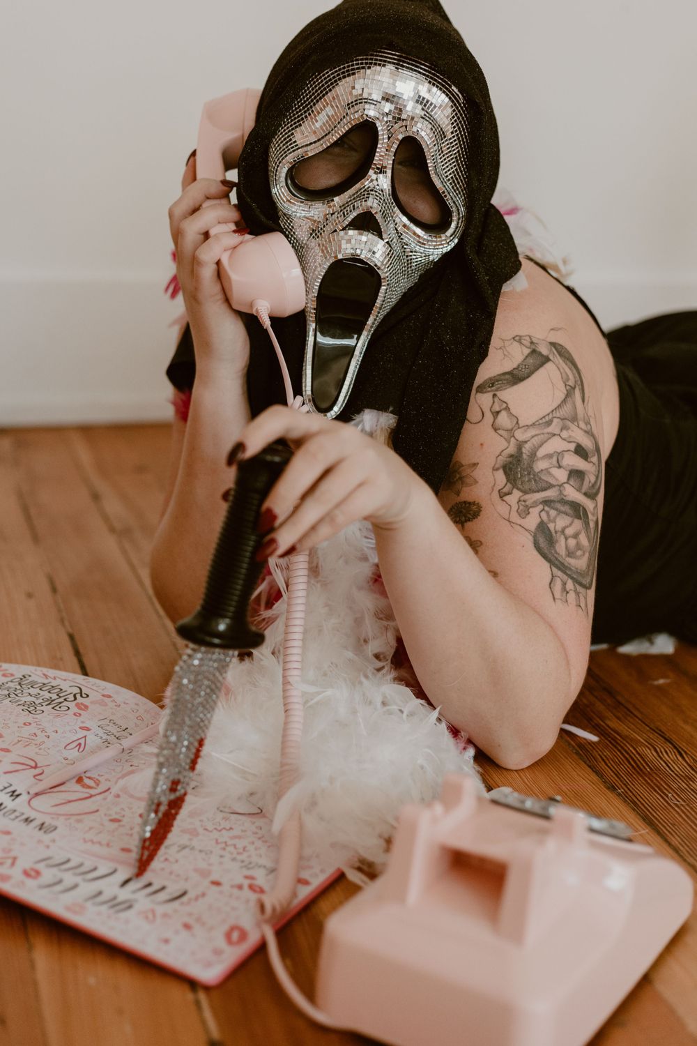 Person wearing sparkly Ghostface mask lying on floor with pink telephone and knife.