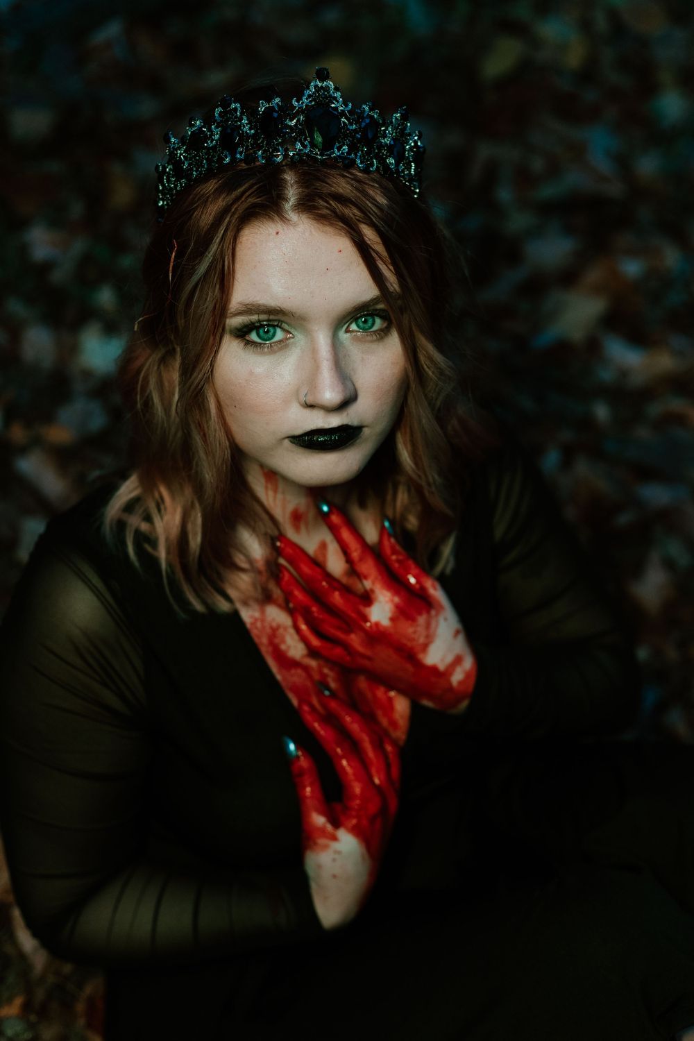 Woman in dark fantasy costume with crown, black lips, and red-painted hands in moody forest.