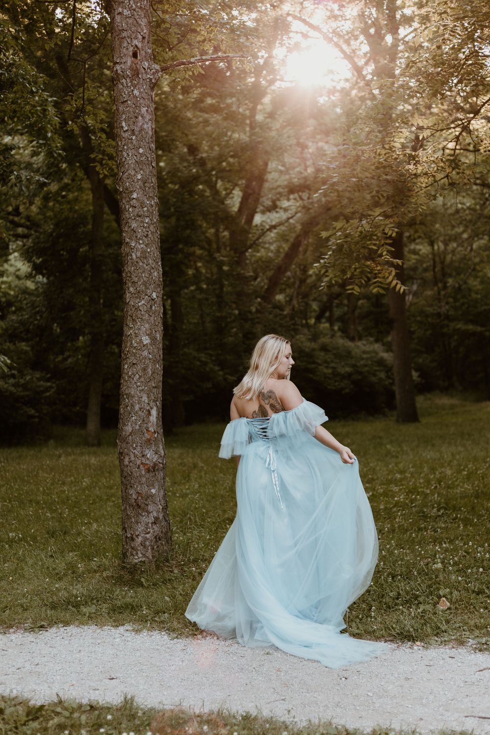 Pregnant woman in flowing light blue off-shoulder gown twirling in a sunlit forest.