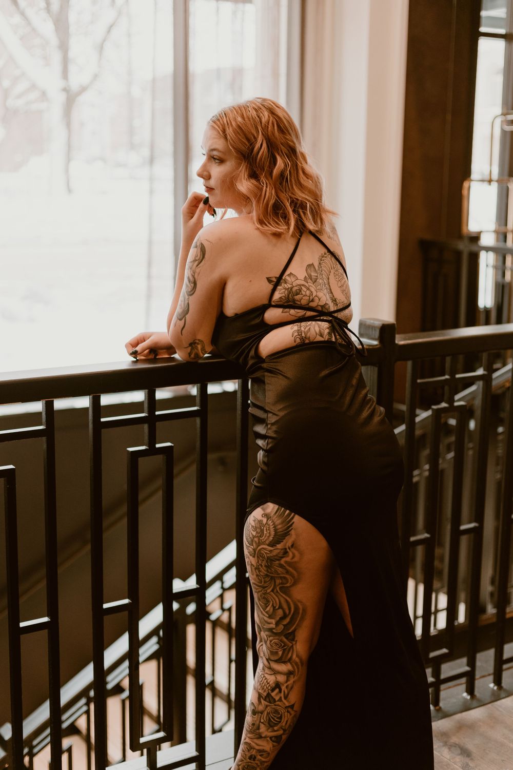 Tattooed redhead woman in elegant black slit gown leaning on staircase railing indoors.