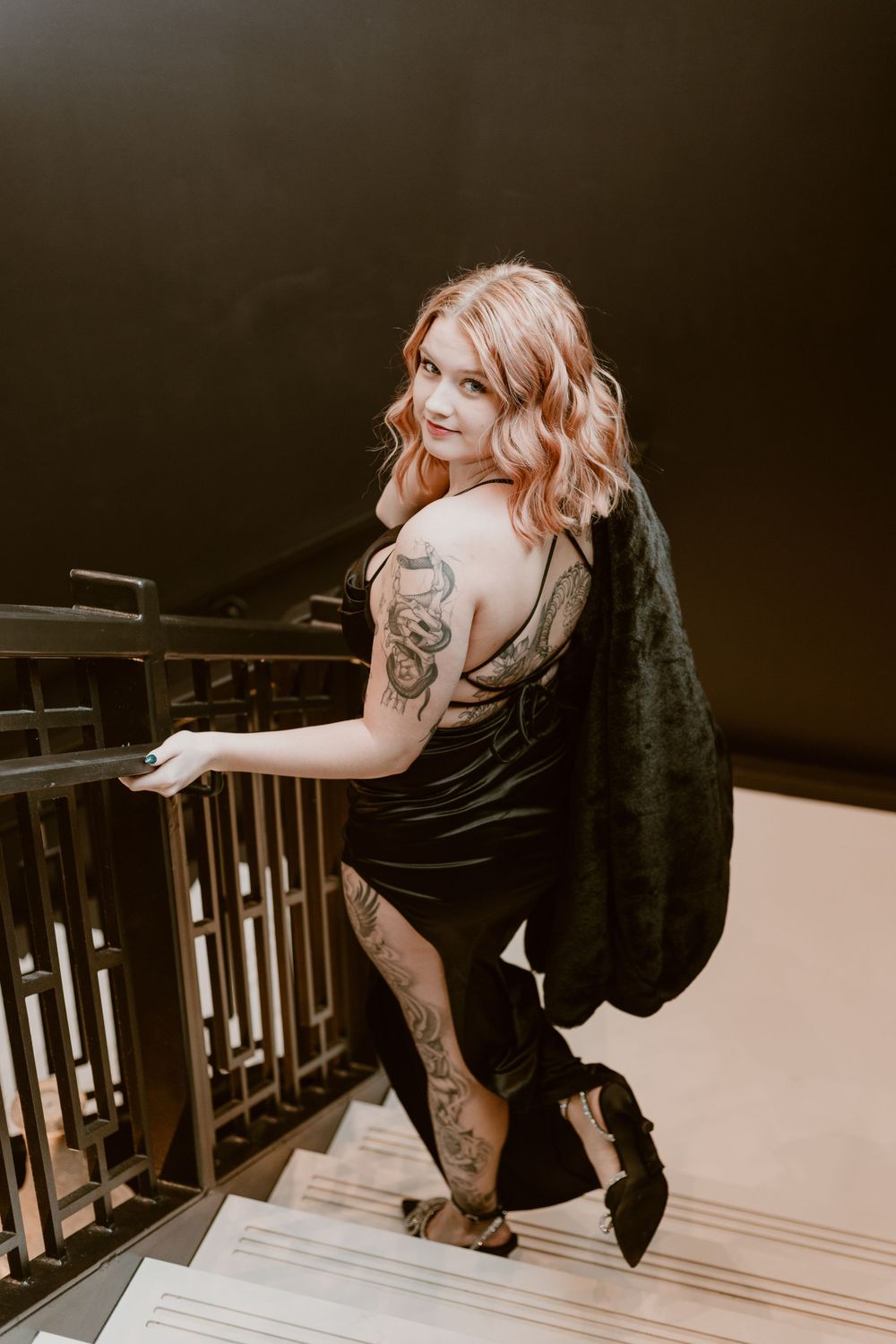 Tattooed woman in black dress carrying jacket while ascending stairs, smiling over shoulder.