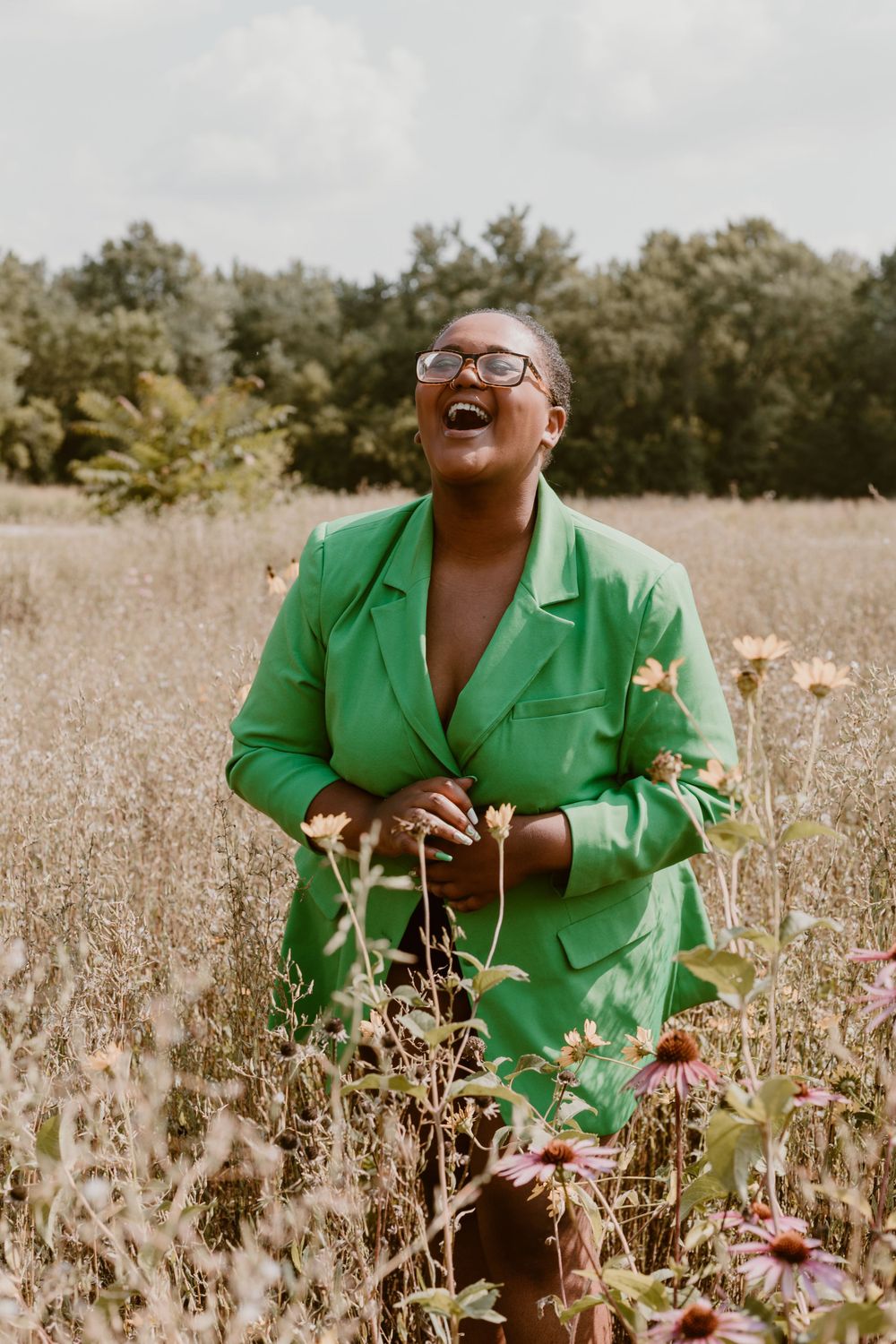 Joyful woman in bright green blazer laughing in a wildflower meadow on a cloudy day.