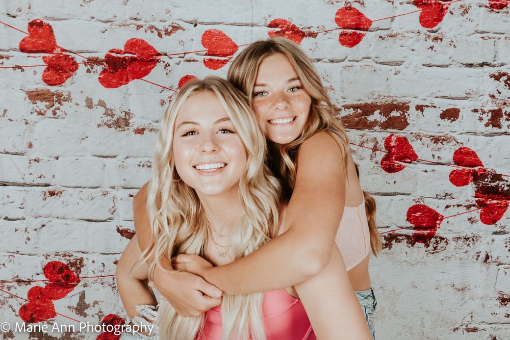 Two smiling teenage girls hugging in front of red heart garland Valentine backdrop.