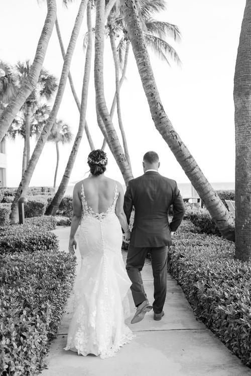 A couple walks down a palm tree-lined path in black and white, with the bride wearing a backless lace gown.