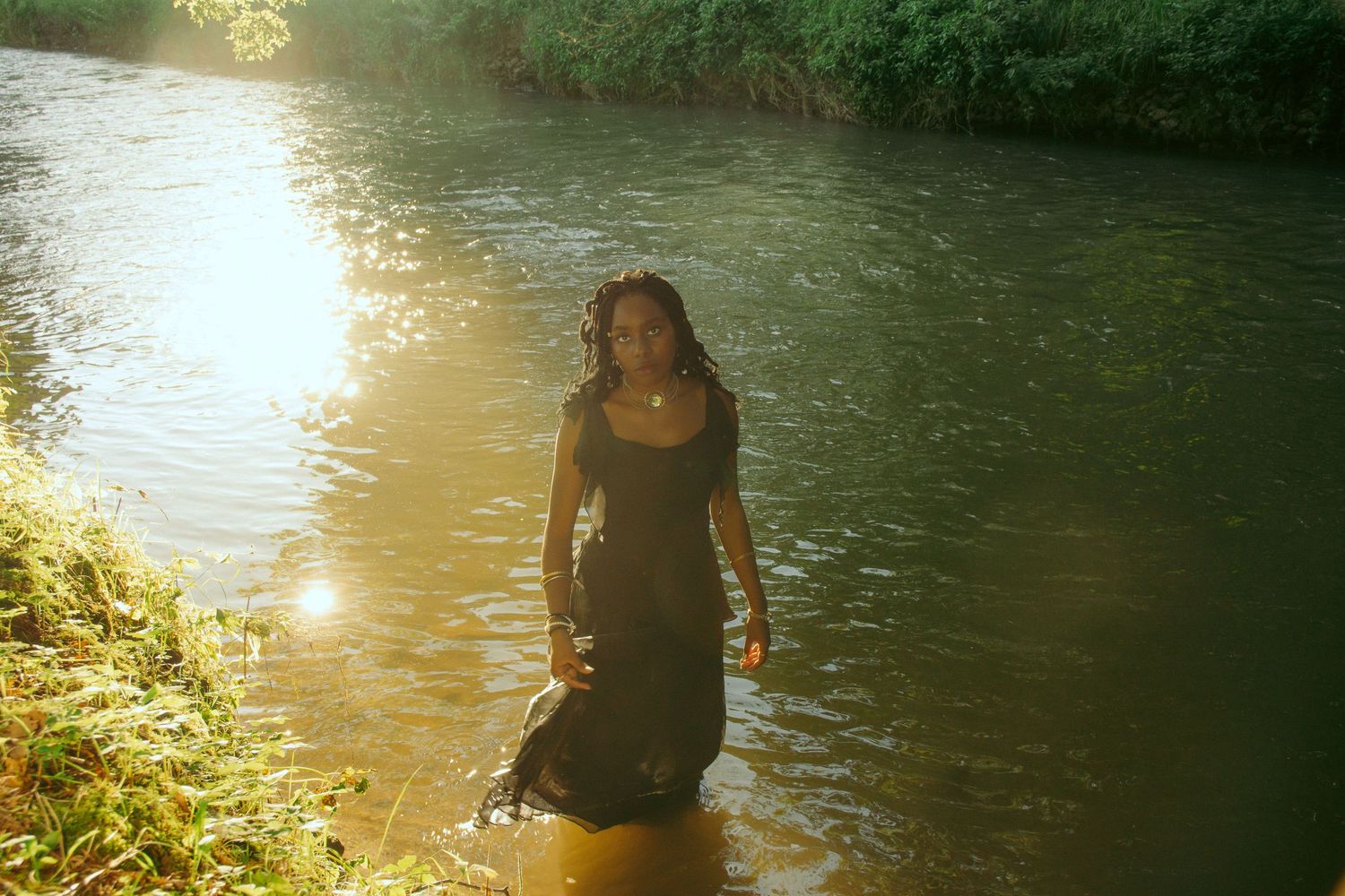 Modest fashion editorial of a woman in a flowing black maxi dress, standing in a river at sunset in Zonhoven, exuding elegance and beauty.