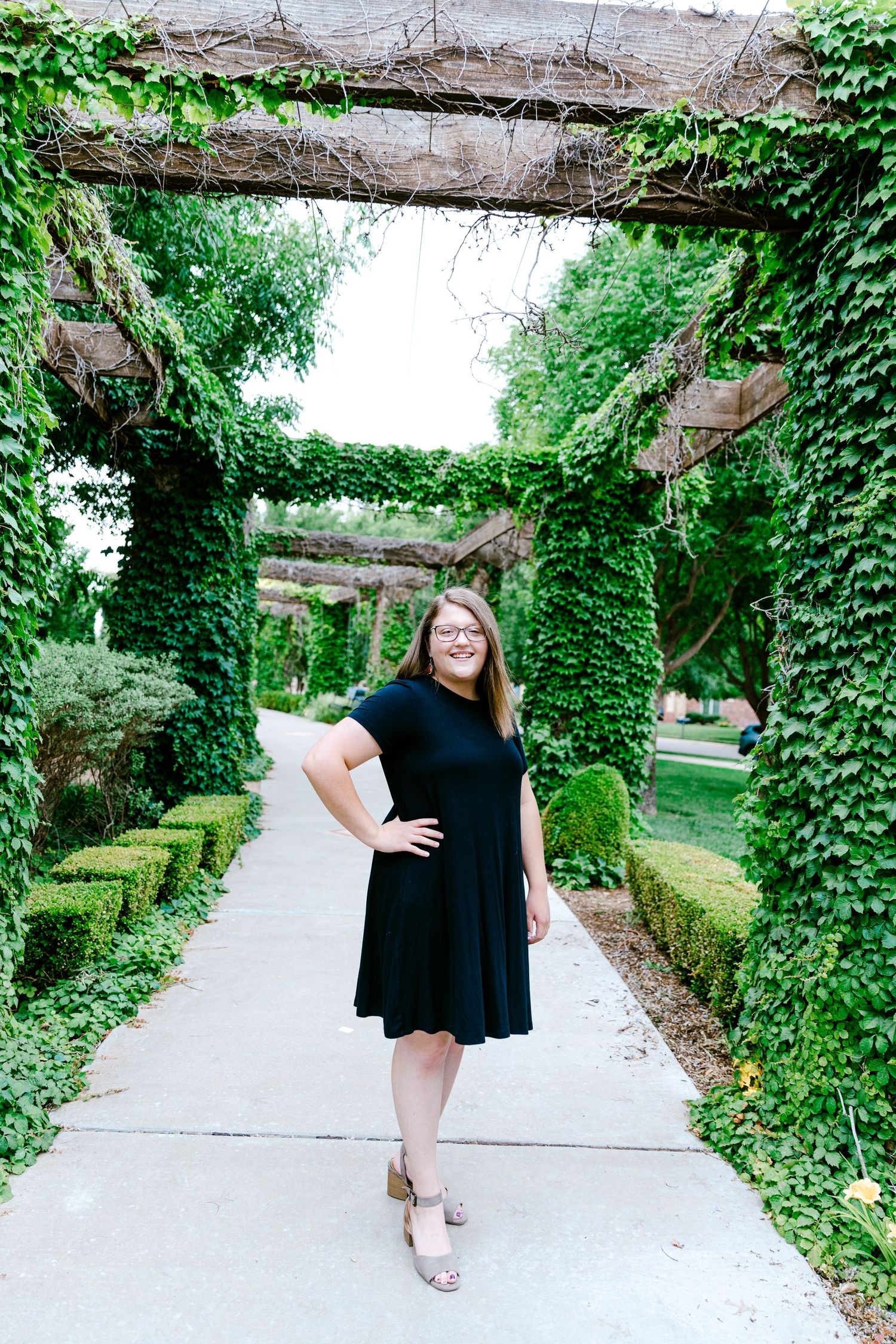 A person in a black dress poses on a garden path beneath an ivy-covered pergola with manicured hedges.