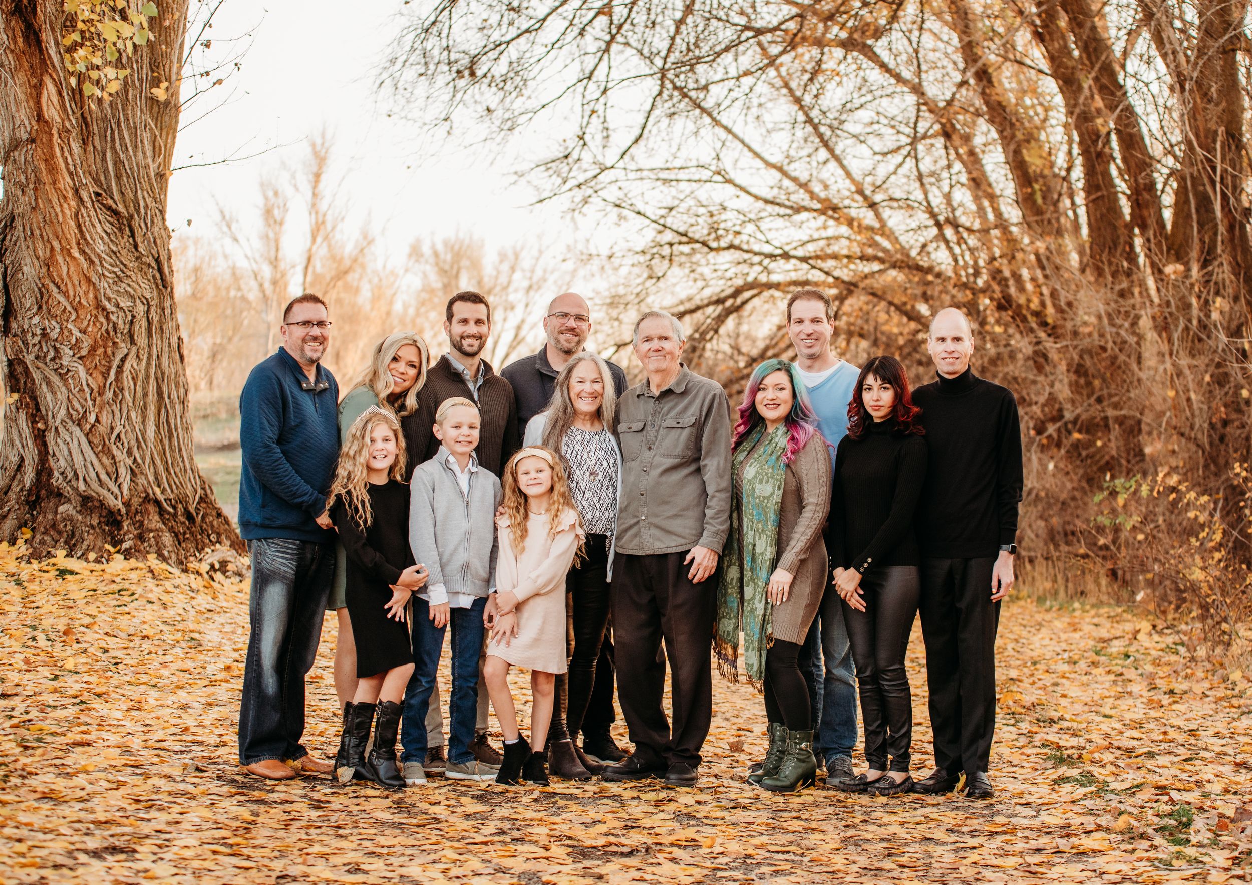 Large family group portrait in casual attire against autumn trees and fallen leaves.