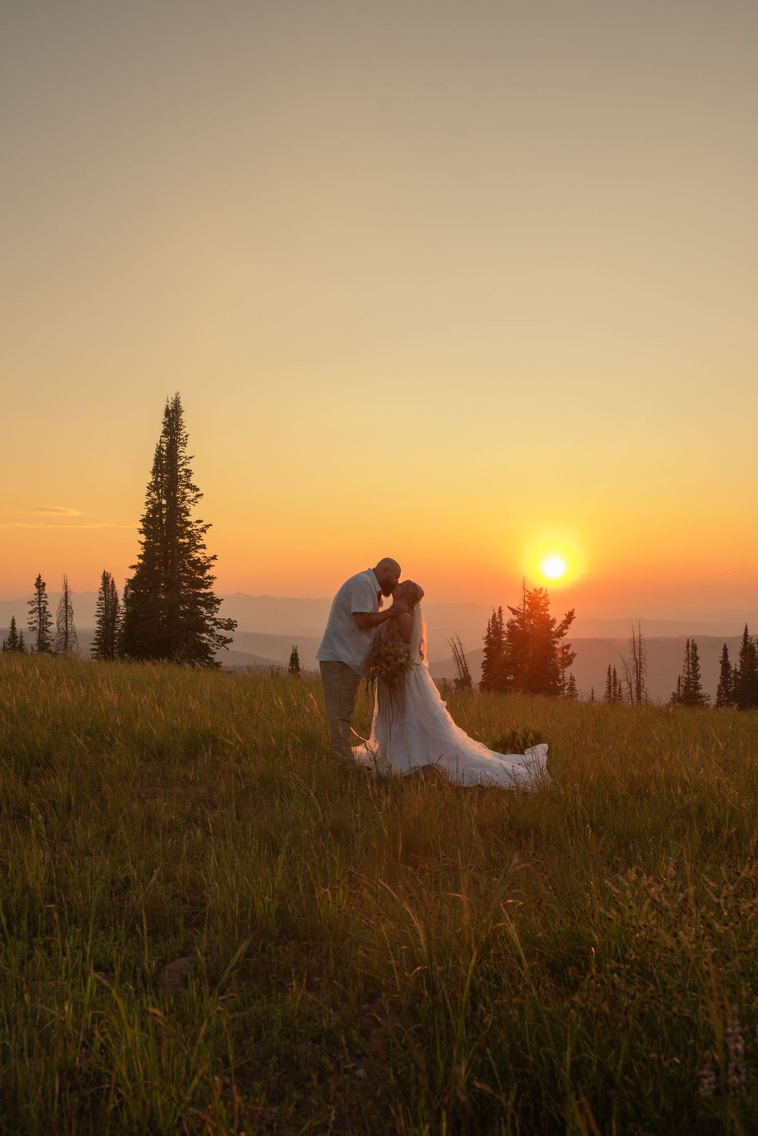 Romantic sunset silhouette of a couple in wedding attire against a mountain backdrop with evergreen trees.