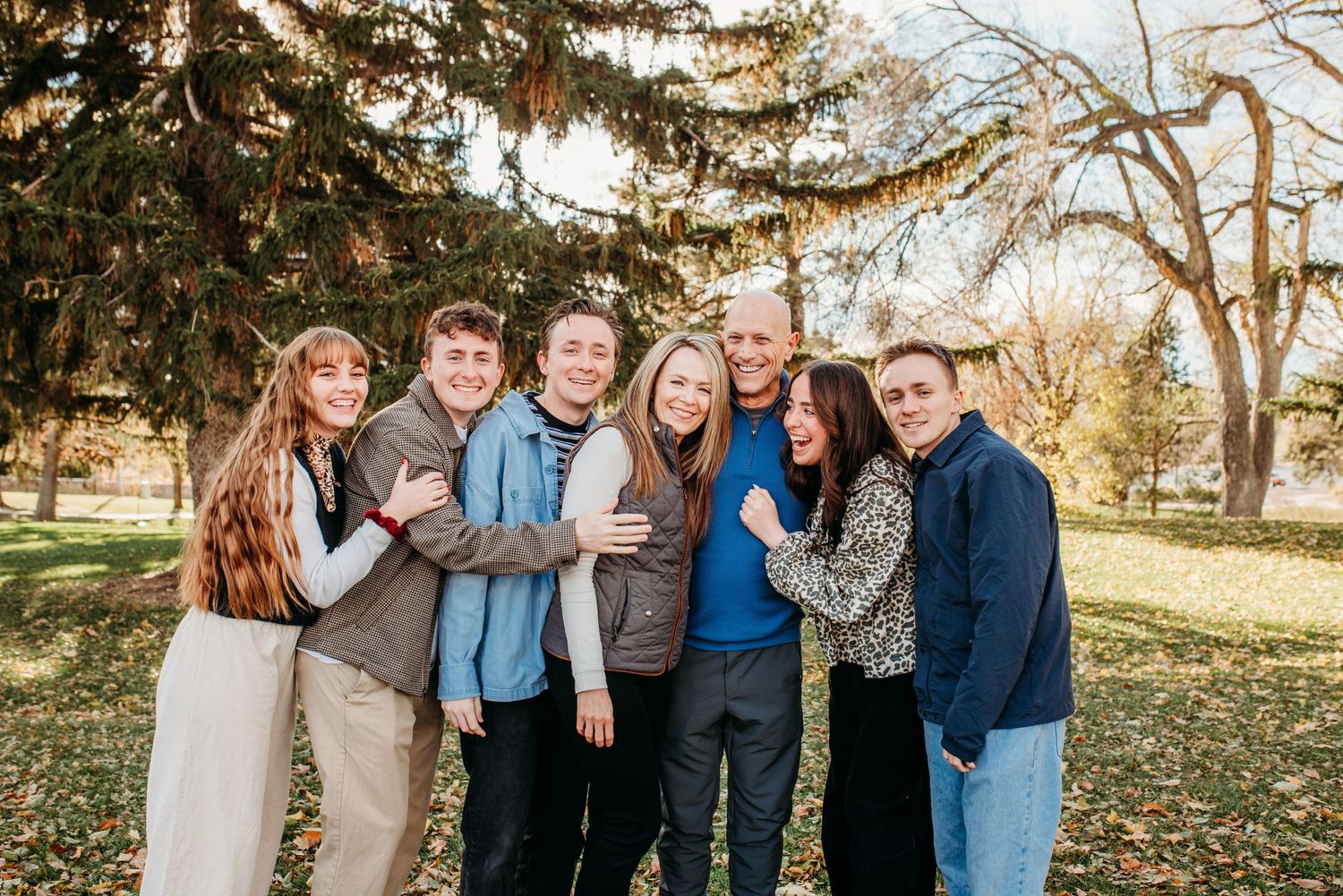 Small group portrait under large evergreen trees in autumn park setting.