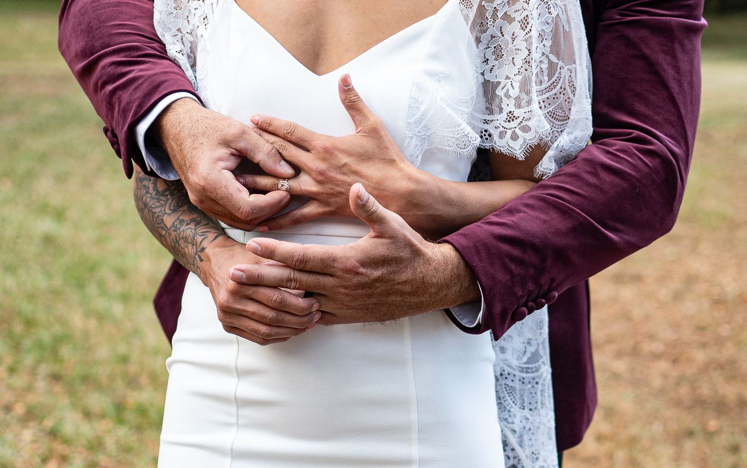 Close-up of bride and groom embracing, showing wedding rings and groom's tattooed arms in outdoor setting.