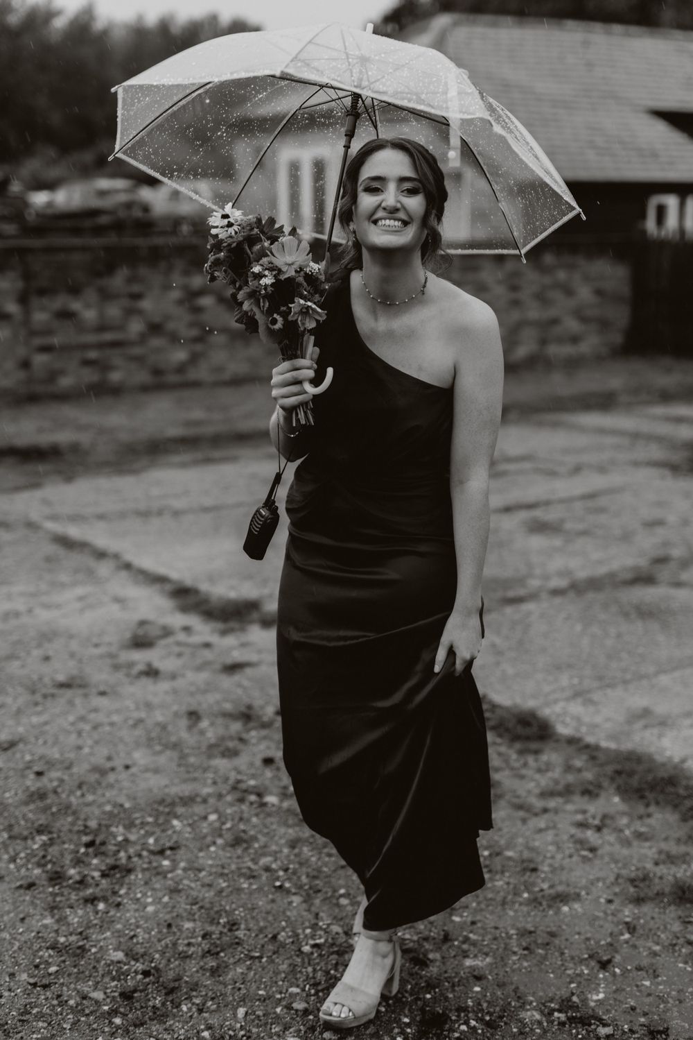 Person in black dress holding clear umbrella poses outdoors in black and white photo.