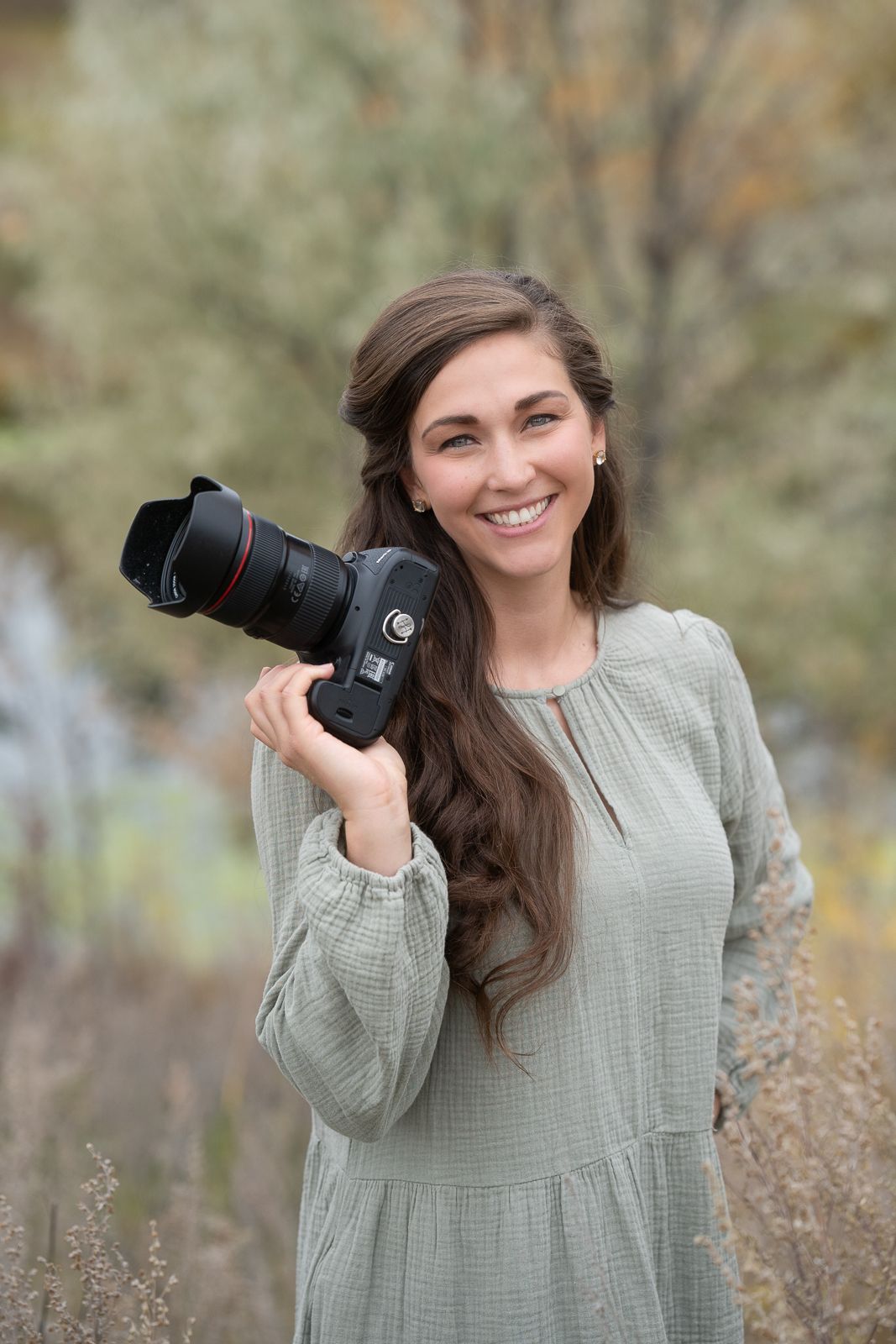 A photographer wearing a green dress holds up a professional camera with a telephoto lens while outdoors in nature.