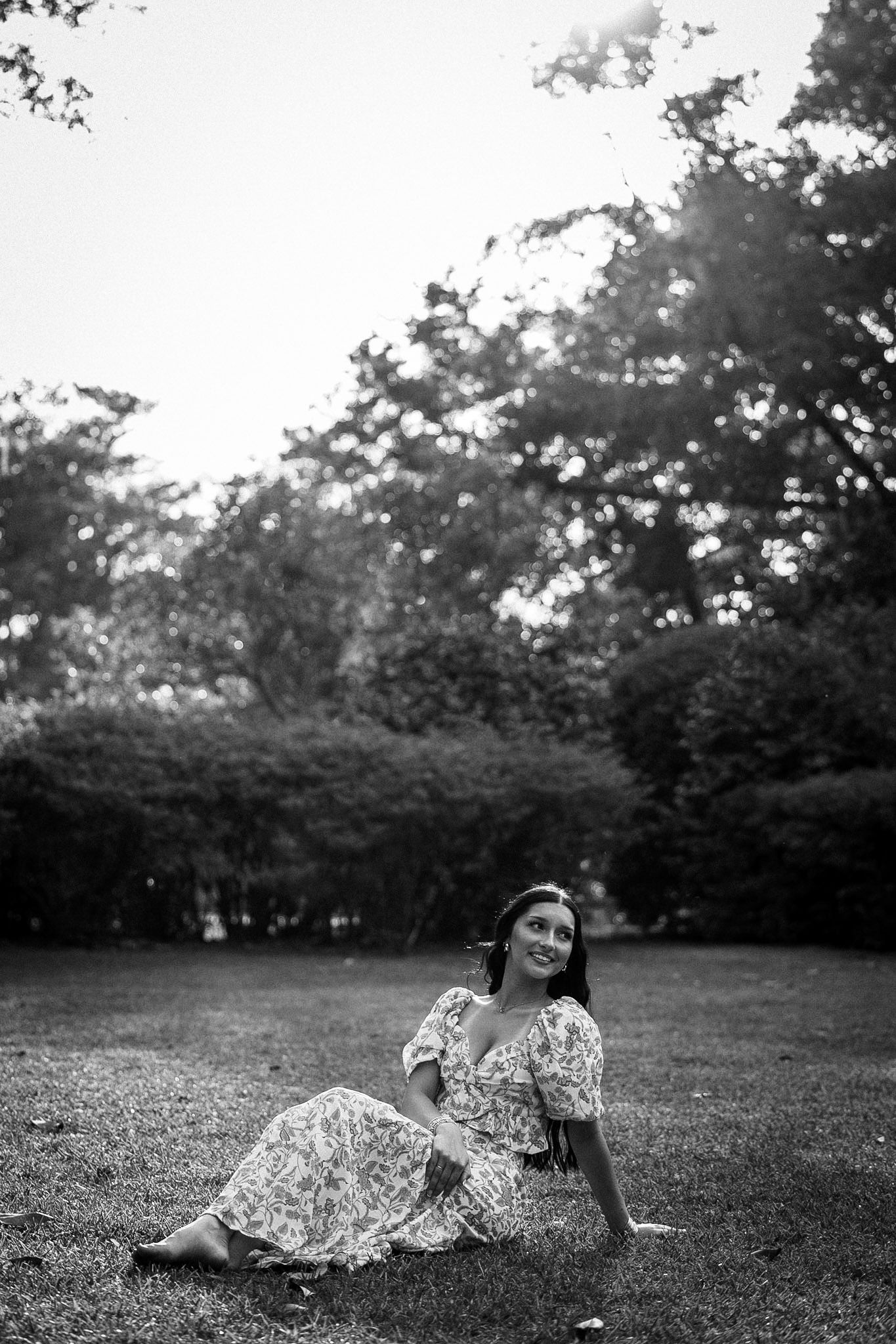 A woman in a floral dress sits gracefully on grass surrounded by trees in a black and white portrait.