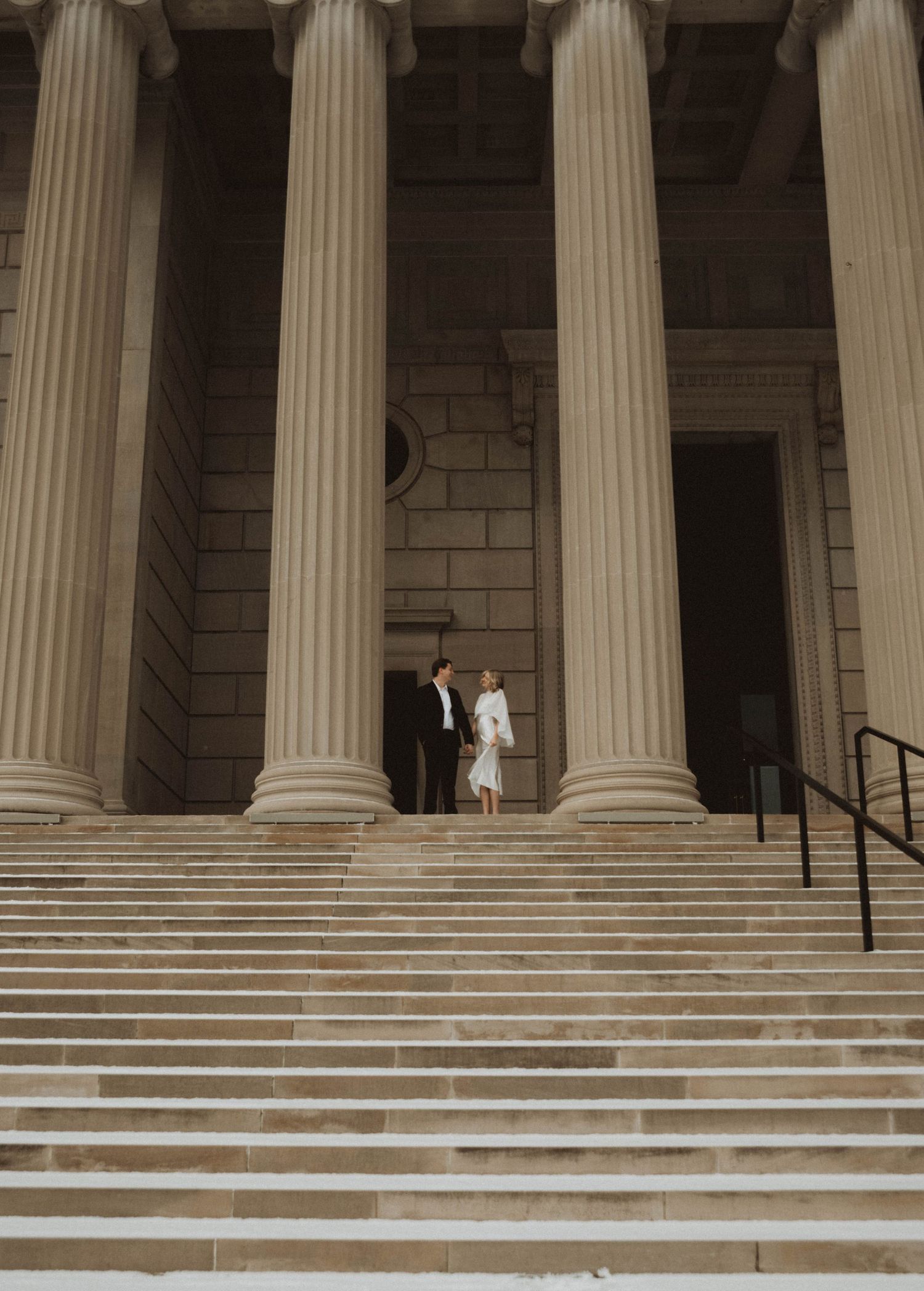 Couple in formal wedding attire standing on grand stone steps of a neoclassical building with tall columns.