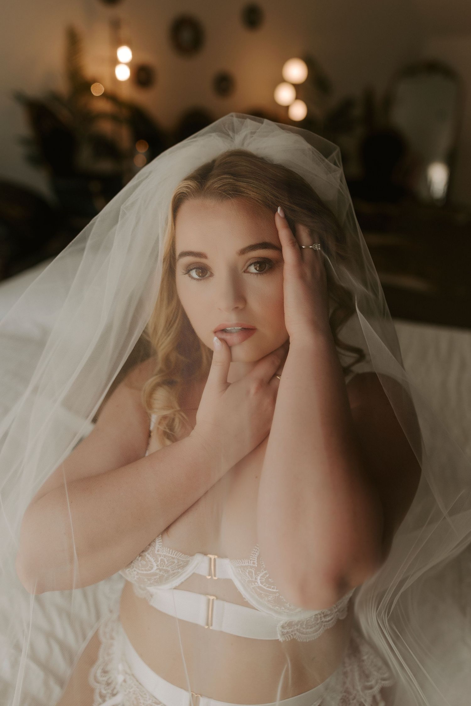 Blonde bride wearing white lace lingerie and veil poses elegantly on bed in softly lit romantic boudoir setting.