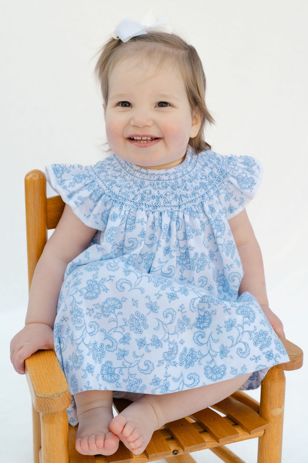 Smiling toddler girl in blue floral smocked dress with white bow sitting on wooden chair against white background.