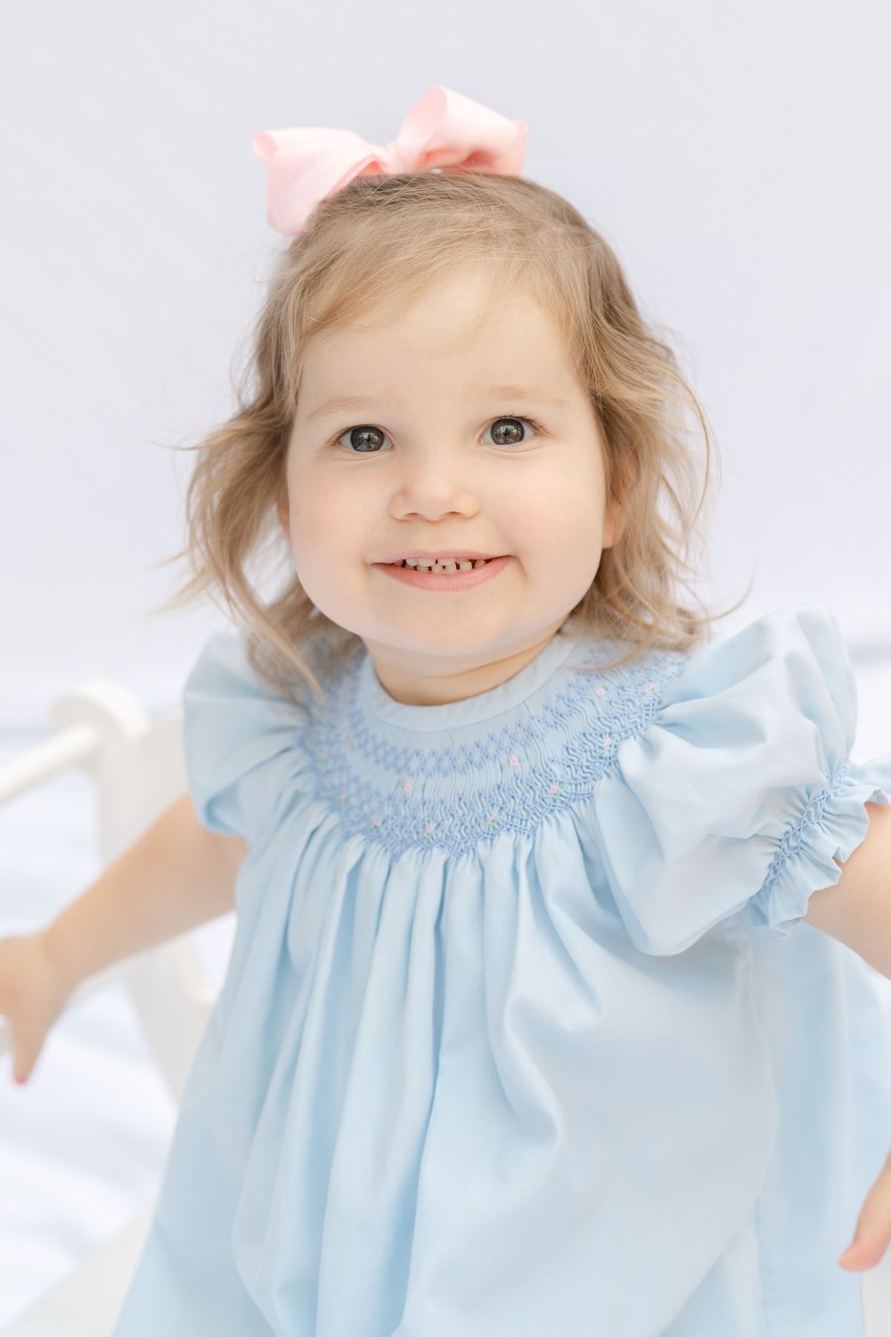 Smiling toddler girl in a light blue smocked dress with a pink bow, posing against a white background.