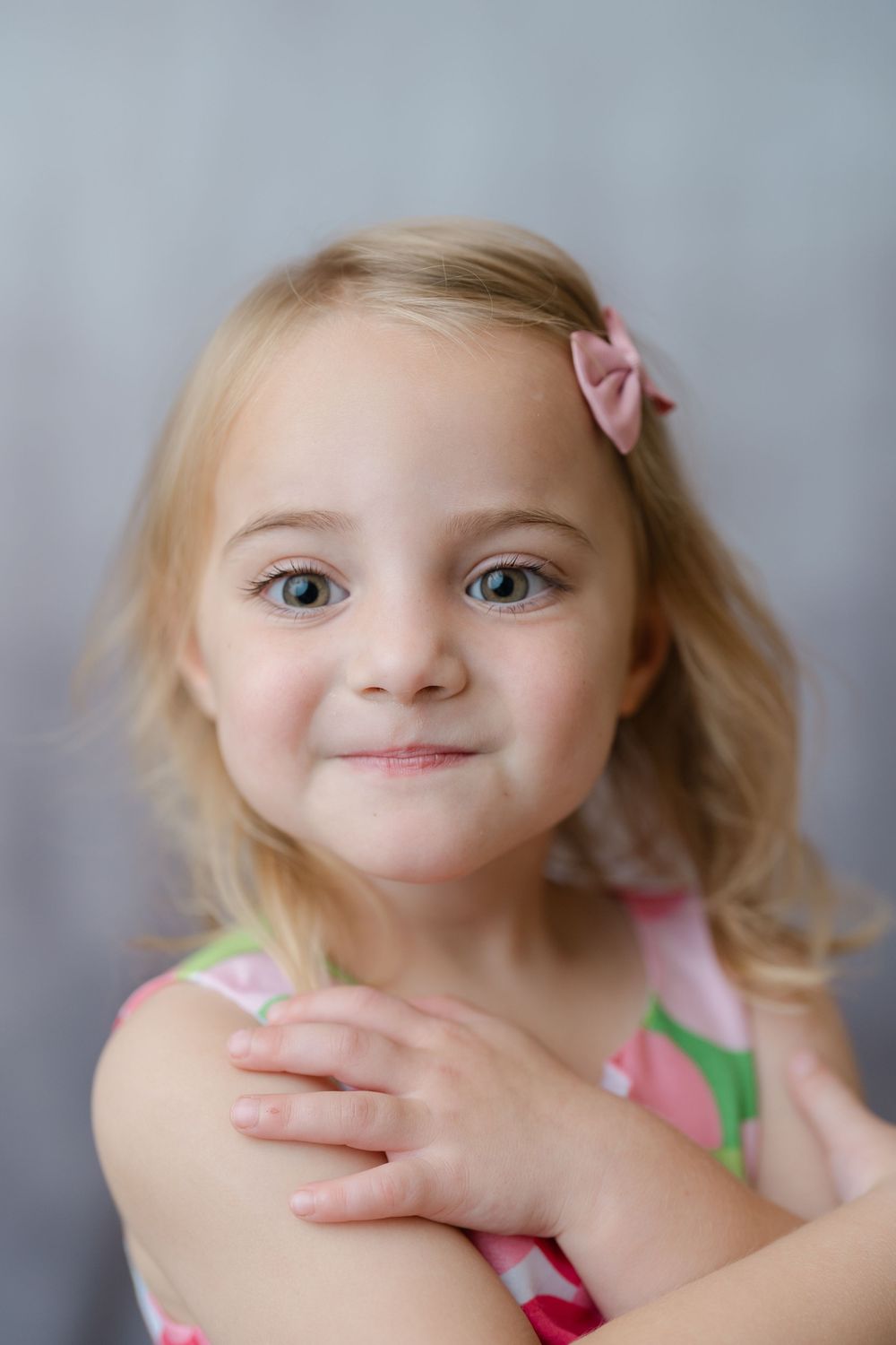 A young blonde girl with blue eyes wearing a pink floral dress and bow hair clip smiles sweetly at the camera.