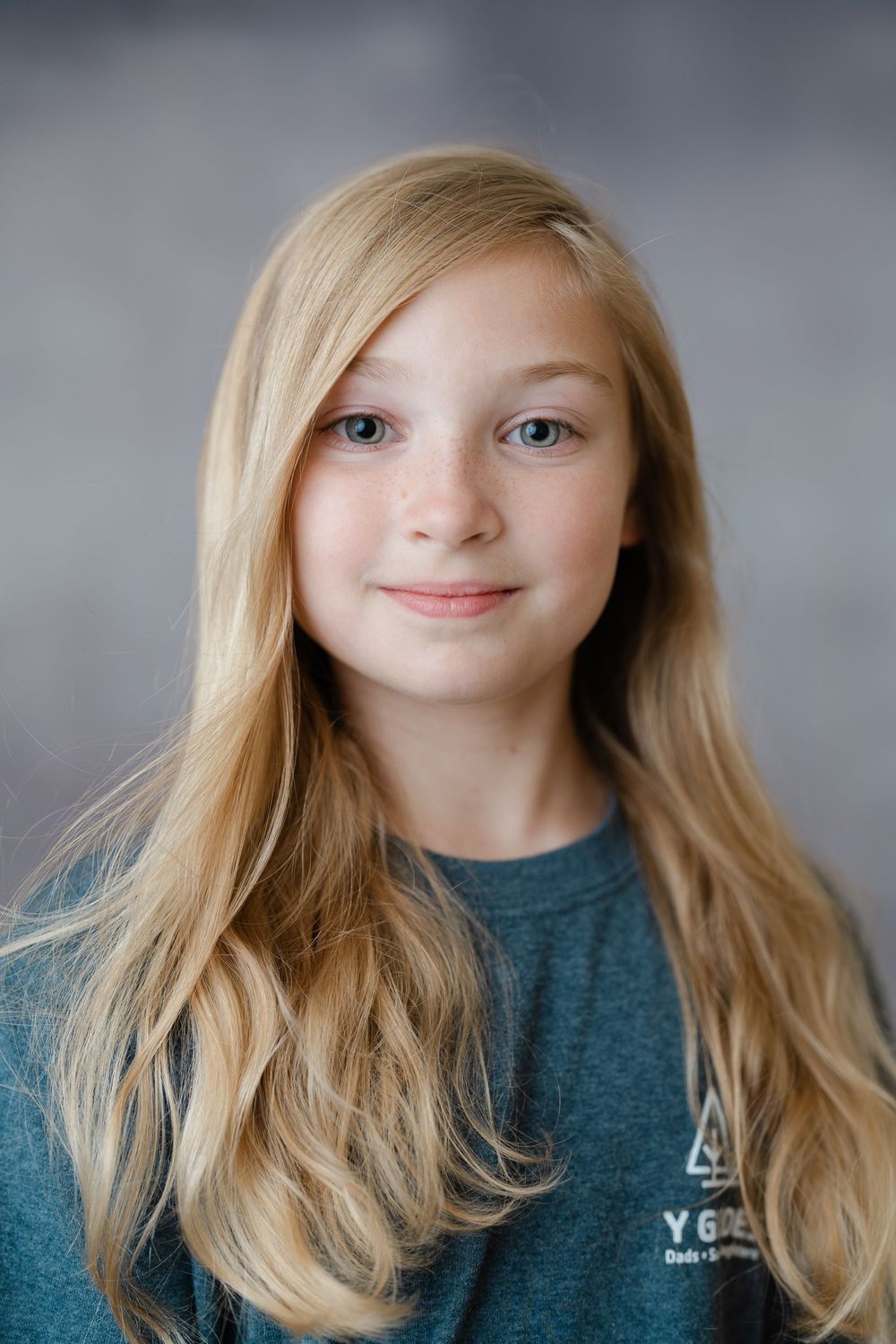 Young blonde girl with long hair and freckles smiling, wearing a teal shirt, portrait photo.