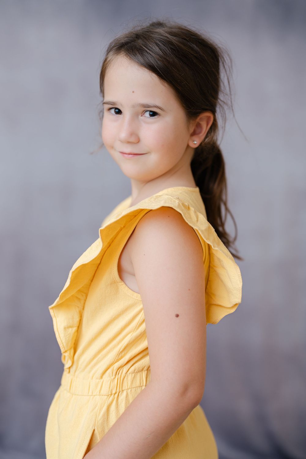 A young girl in a yellow sleeveless dress poses against a gray background with a gentle smile.