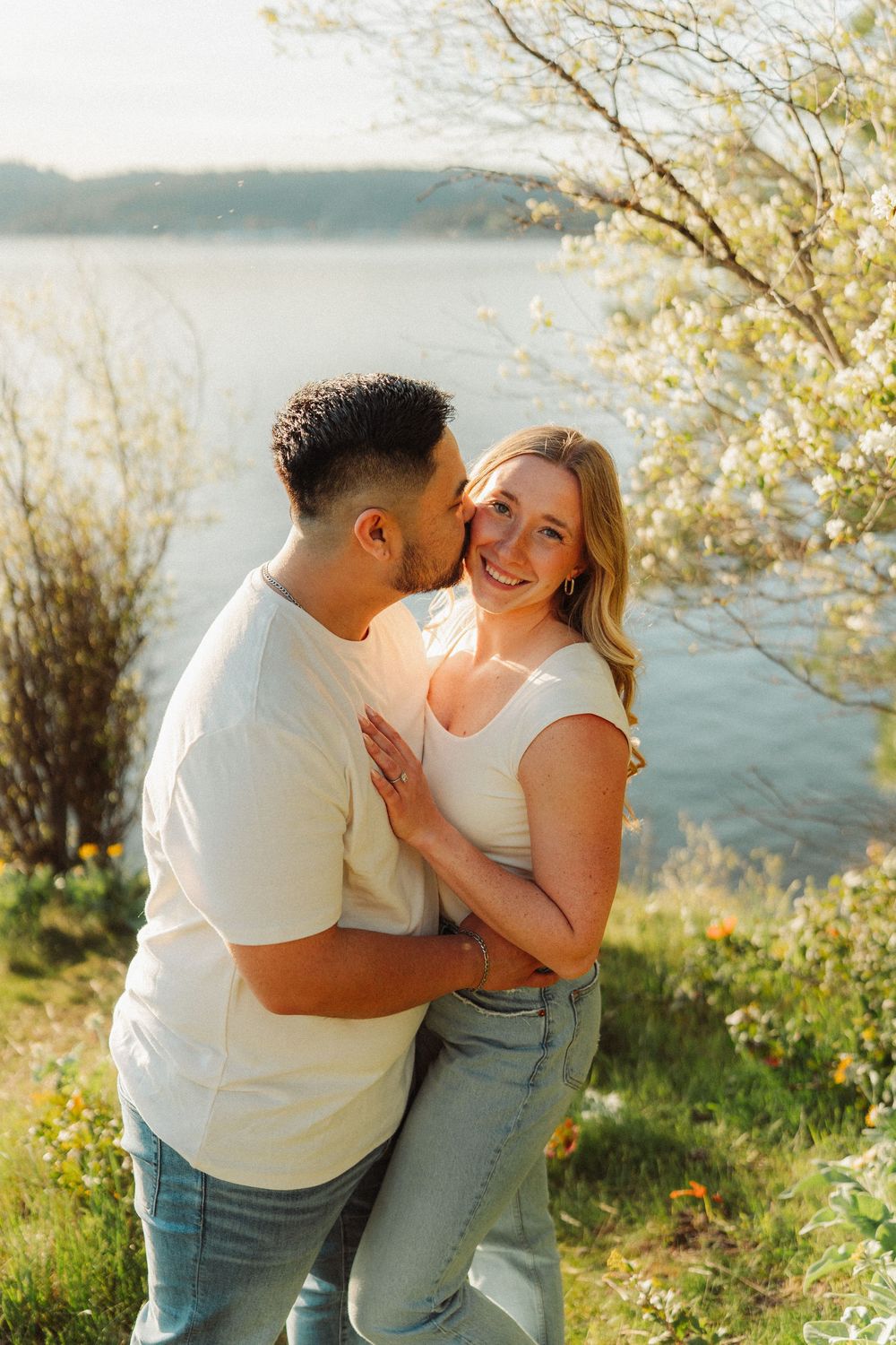 Couple embracing by a lake at golden hour, man kissing woman's cheek.