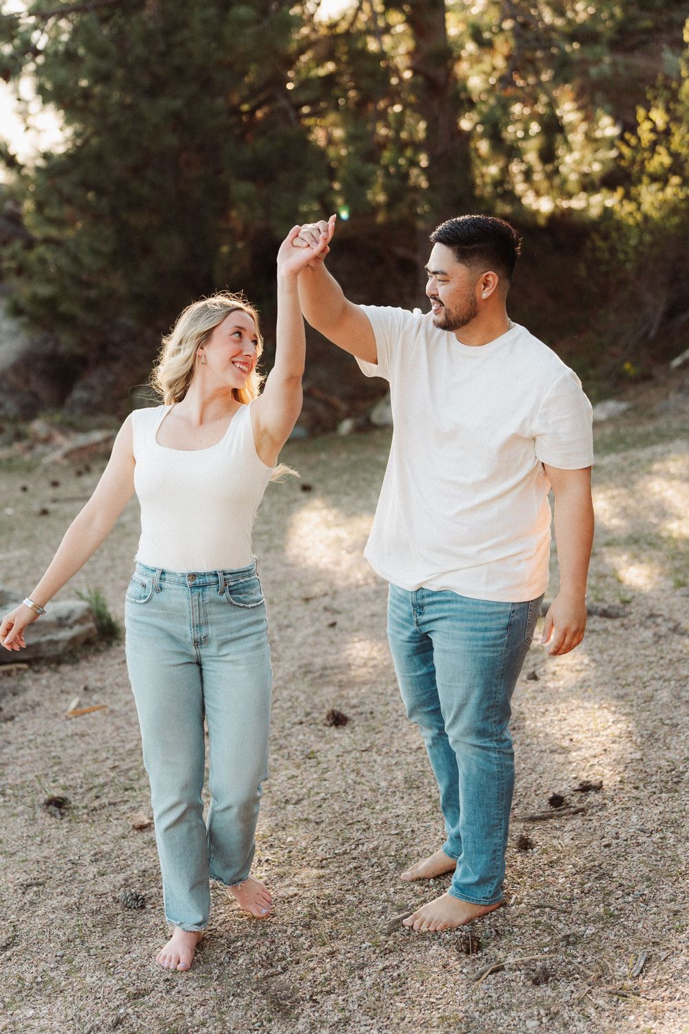 Happy couple dancing barefoot on sandy beach, wearing matching white tops and jeans, holding hands in golden sunlight.