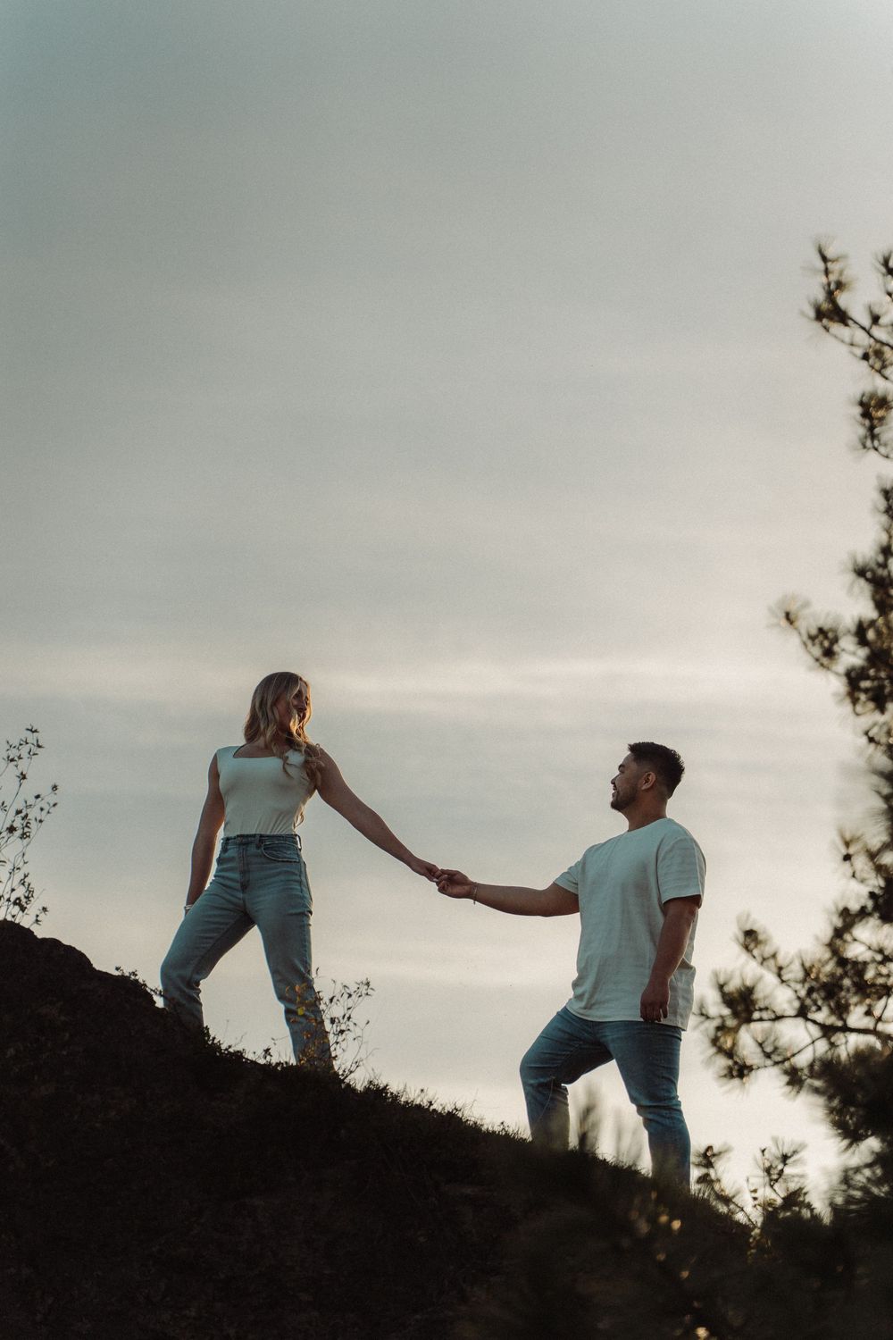 A couple holds hands while climbing a rocky hill at dusk, dressed in white tops and jeans against a cloudy sky.