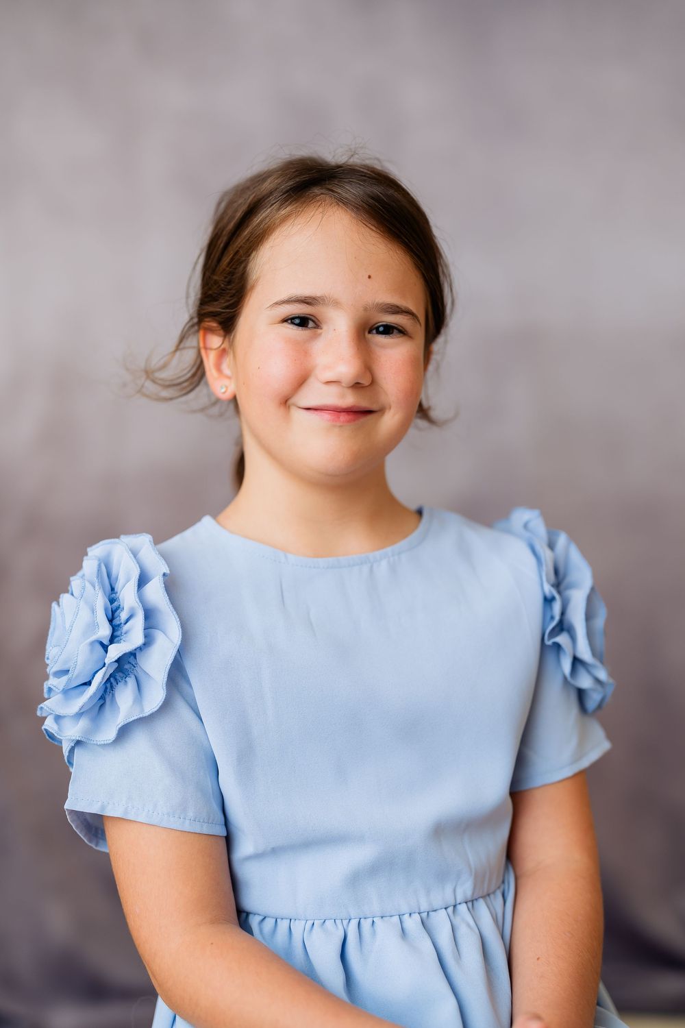 Young girl in a light blue dress with floral sleeves smiling at the camera in a studio portrait.