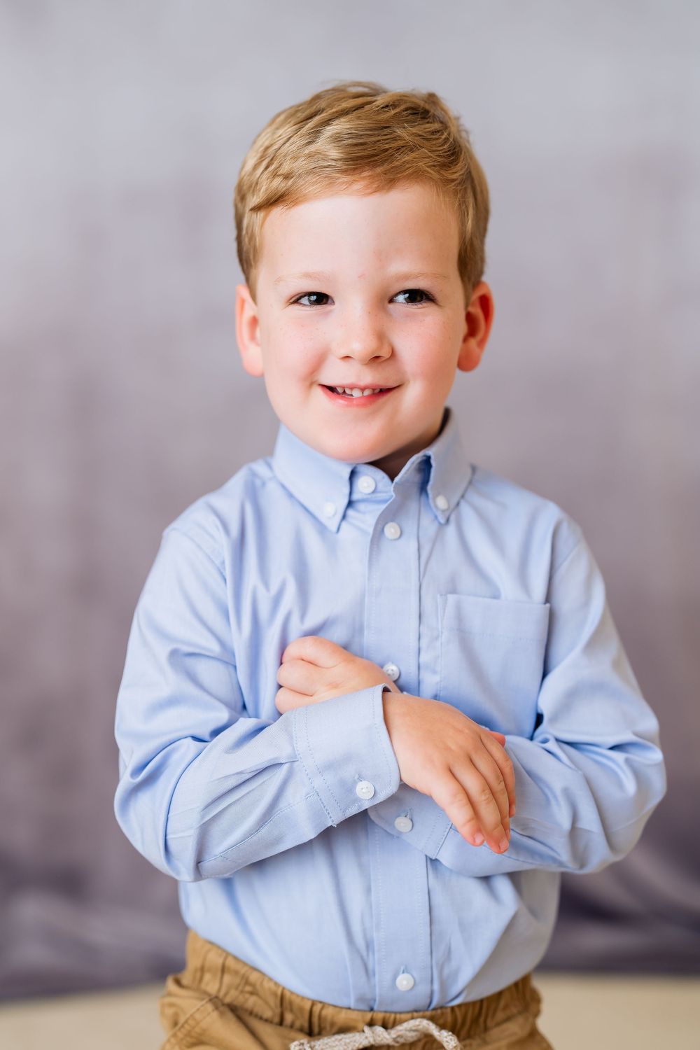 Smiling young boy in a light blue button-up shirt with arms crossed, posing for a portrait photo.