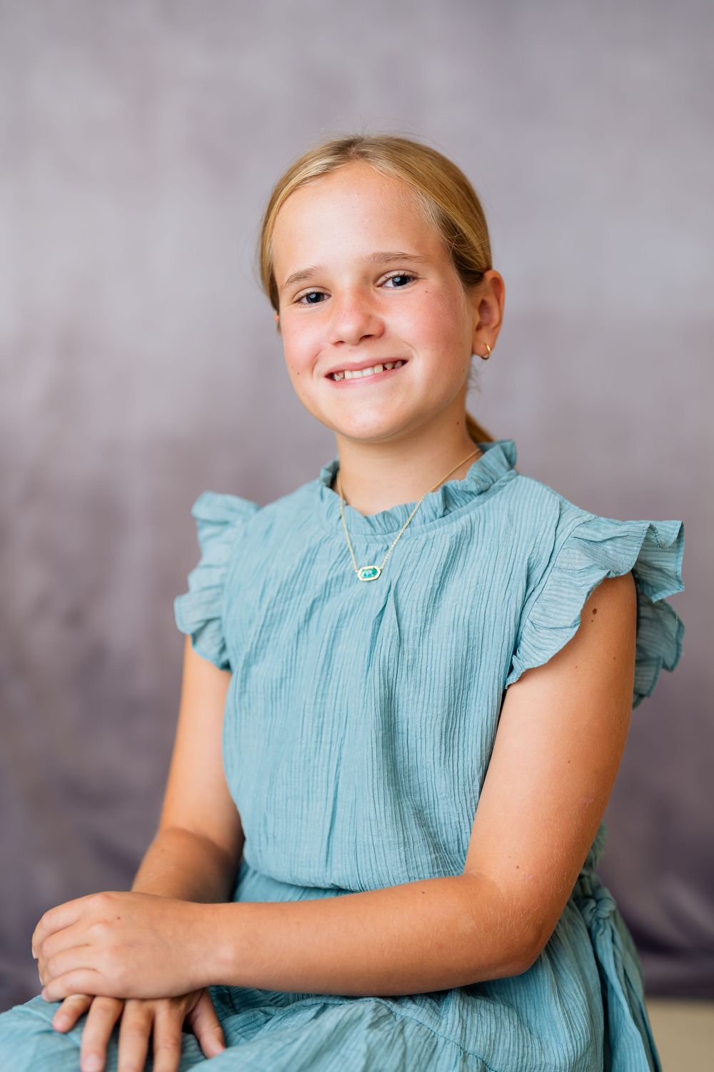Smiling young girl in teal ruffle dress with necklace, posed for a portrait against gray studio background.