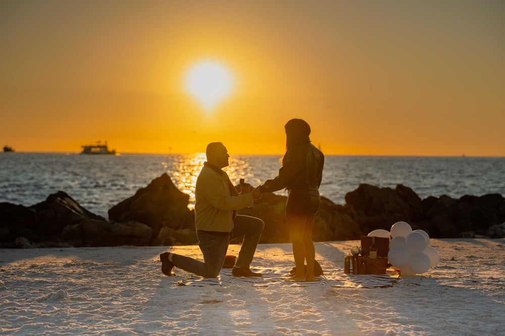 Silhouette of a marriage proposal on a snowy beach at golden sunset with ocean waves and rocks in background.