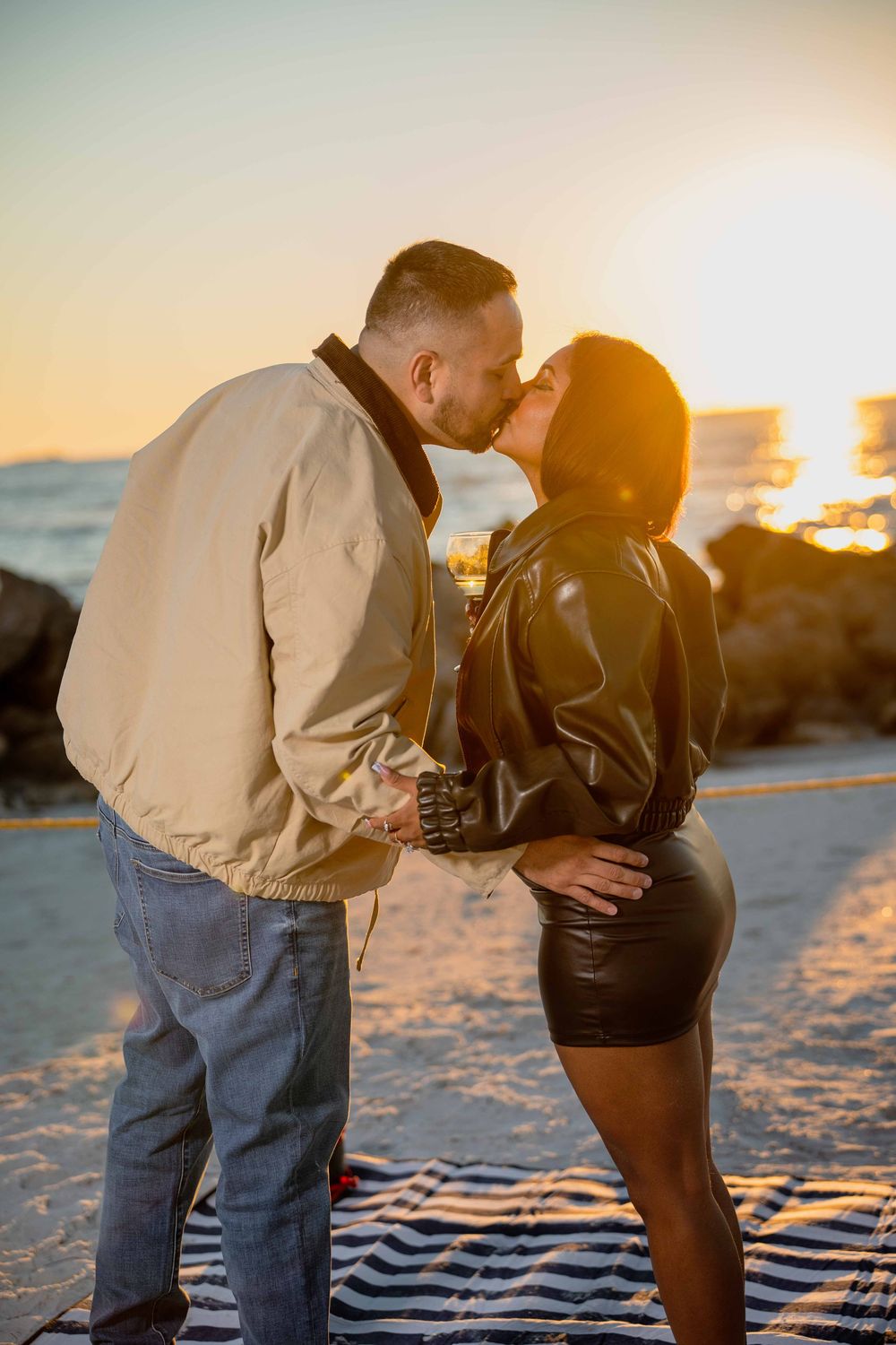Couple sharing a romantic kiss on the beach at golden sunset, wearing leather and bomber jackets on sandy shore.