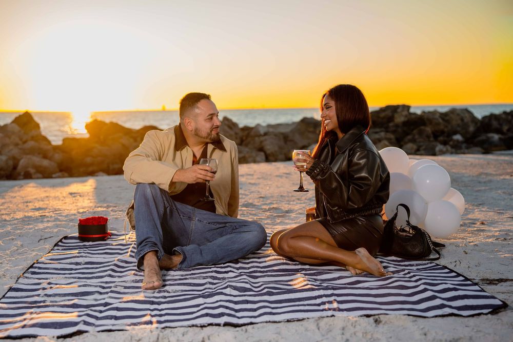 Couple enjoying a romantic beach picnic at sunset, toasting with wine glasses on a striped blanket near rocks.