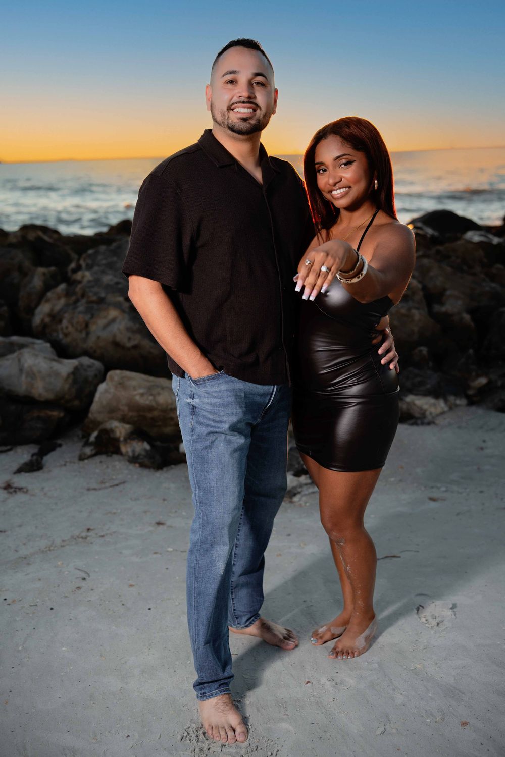 Couple posing on a sandy beach at sunset, woman showing engagement ring, rocks and colorful sky in background.