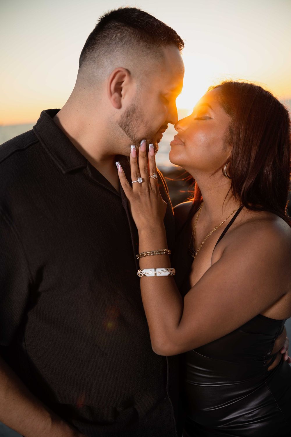 Couple sharing a romantic sunset moment, woman displaying engagement ring while almost kissing outdoors.