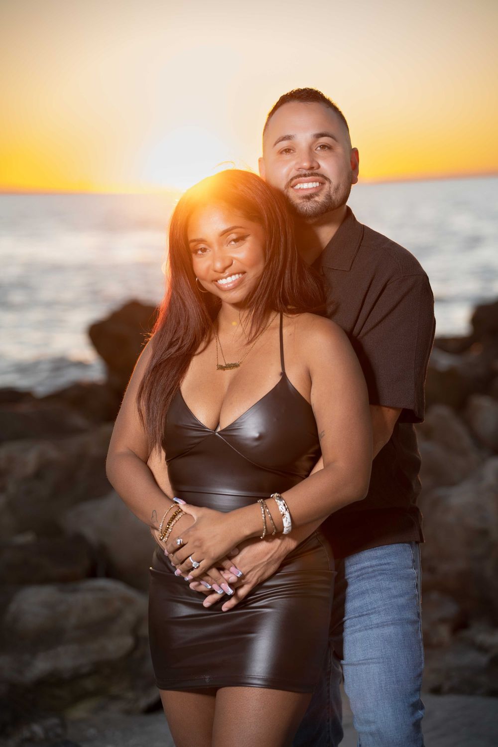 Couple embracing at sunset by rocky ocean shore, woman in black leather dress, man in brown shirt, golden hour backdrop.