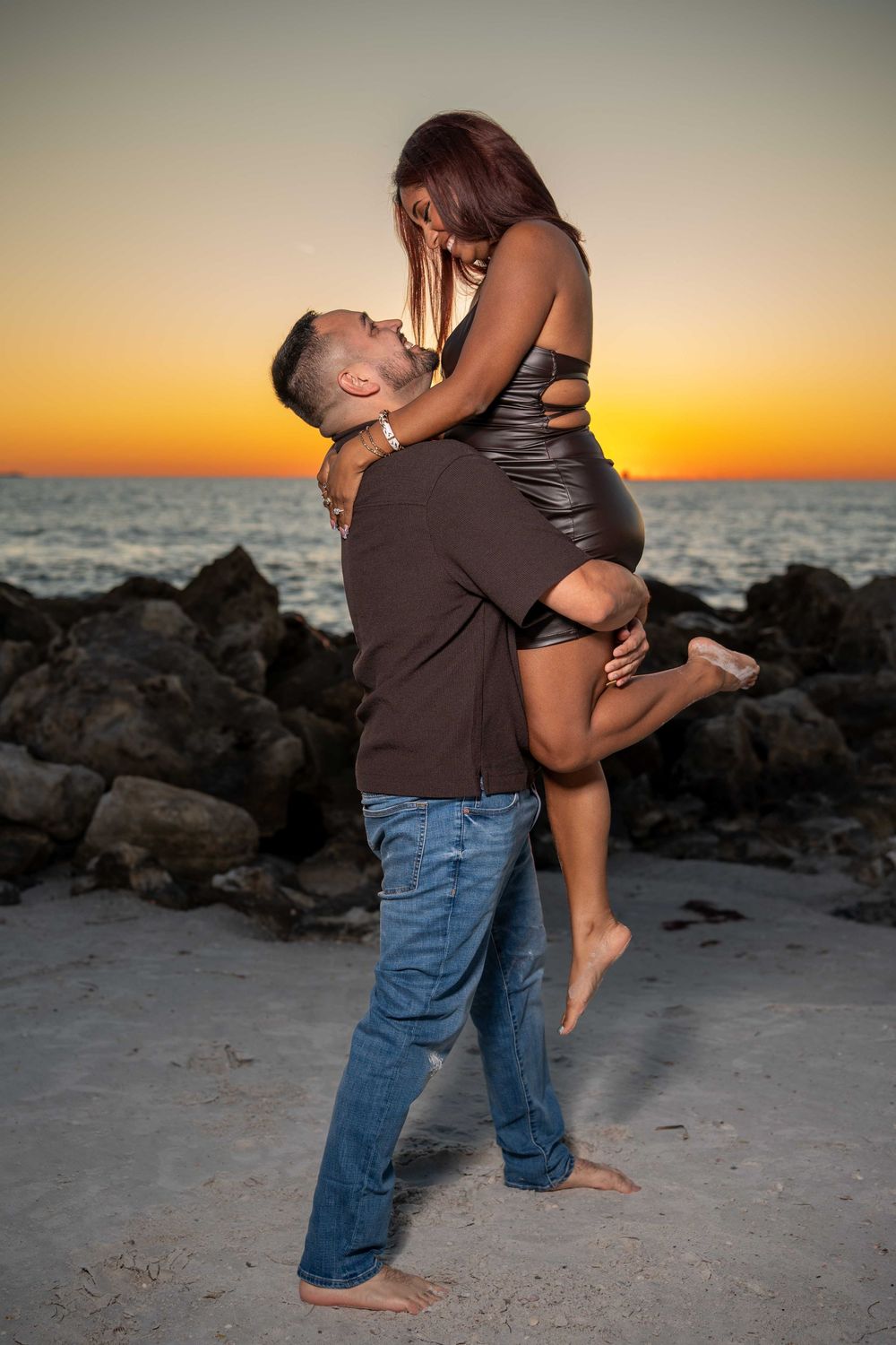 Couple sharing a romantic moment at sunset on a rocky beach, man lifting woman in black dress.