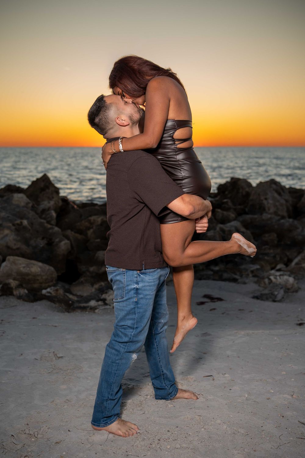 Couple sharing a romantic kiss on a sandy beach at sunset, man lifting woman in black dress near rocky shoreline.