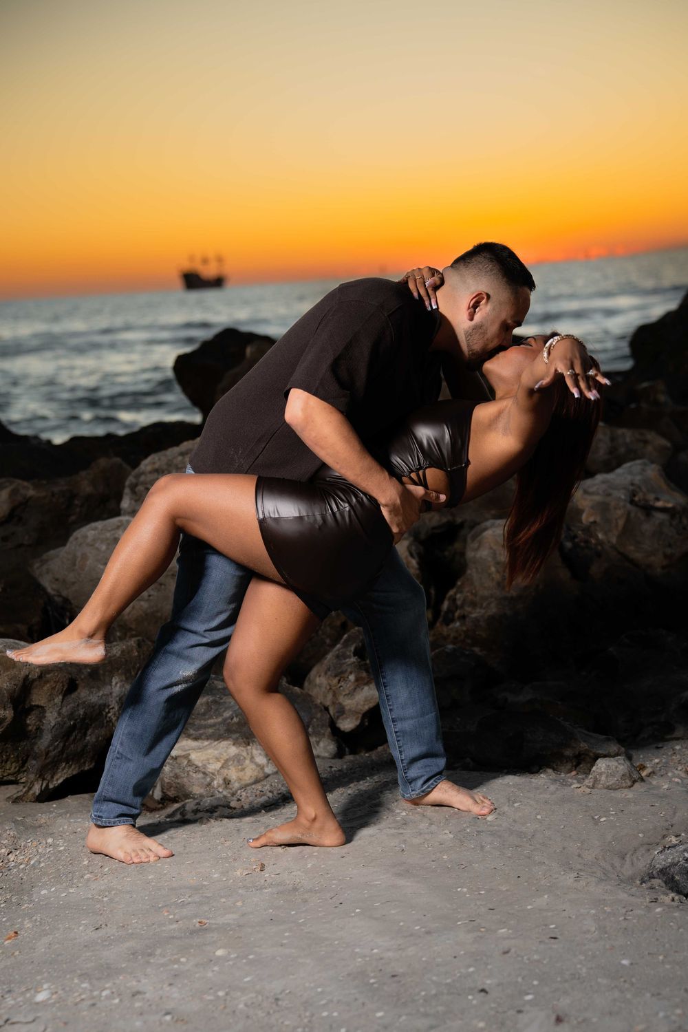 Couple sharing a romantic kiss on a rocky beach at sunset with golden sky and ocean waves in the background.