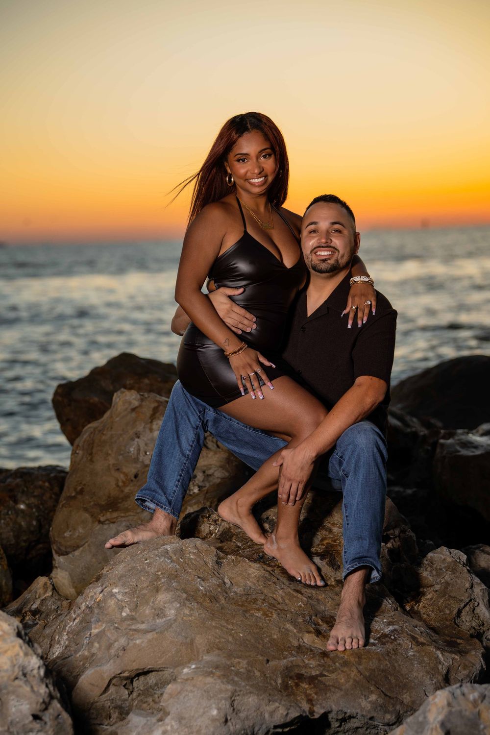 A couple poses on rocky shoreline at sunset, woman in black bodysuit and jeans sitting on mans lap, ocean behind them.