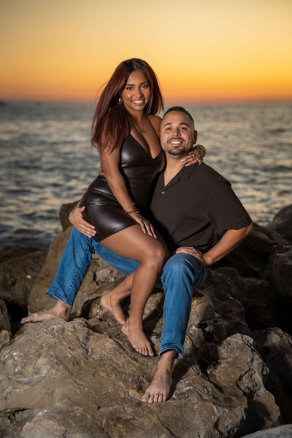 Couple posing on rocks at sunset by the ocean, woman in leather dress sitting on mans lap, golden sky backdrop.
